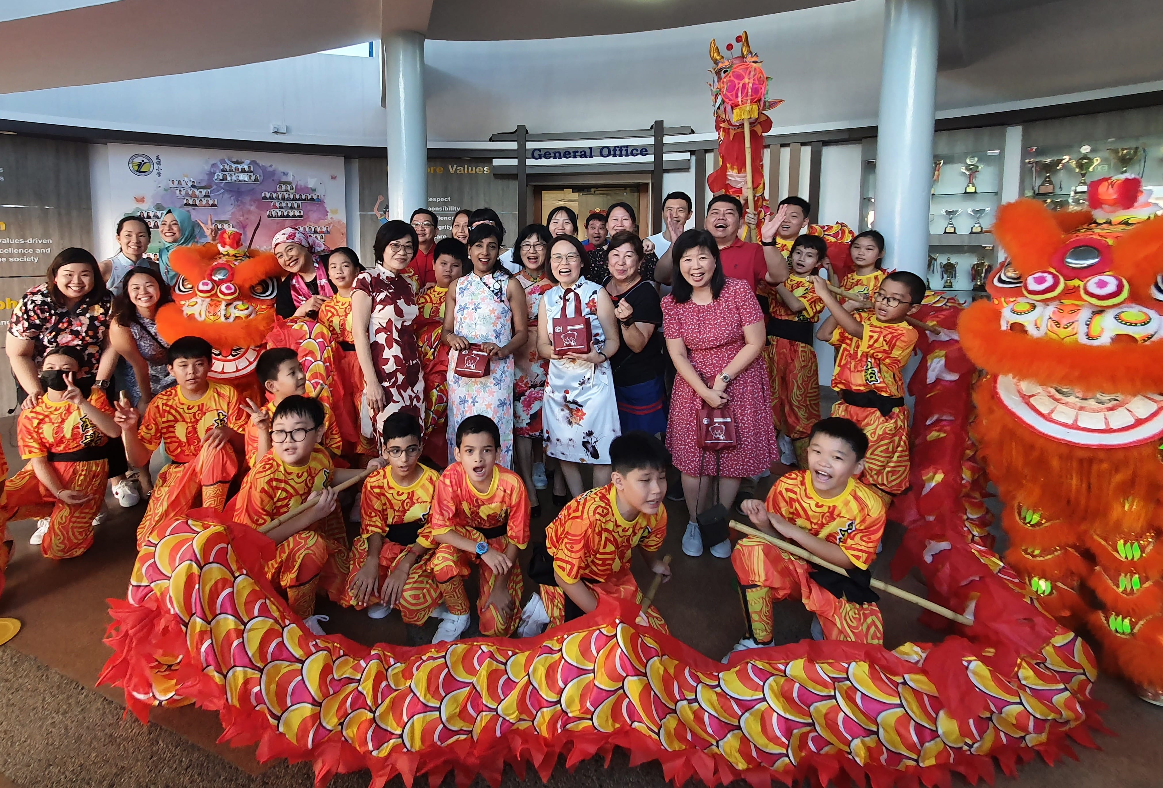 Children in red and yellow costumes pose with adults beside colorful lion and dragon dance props indoors.