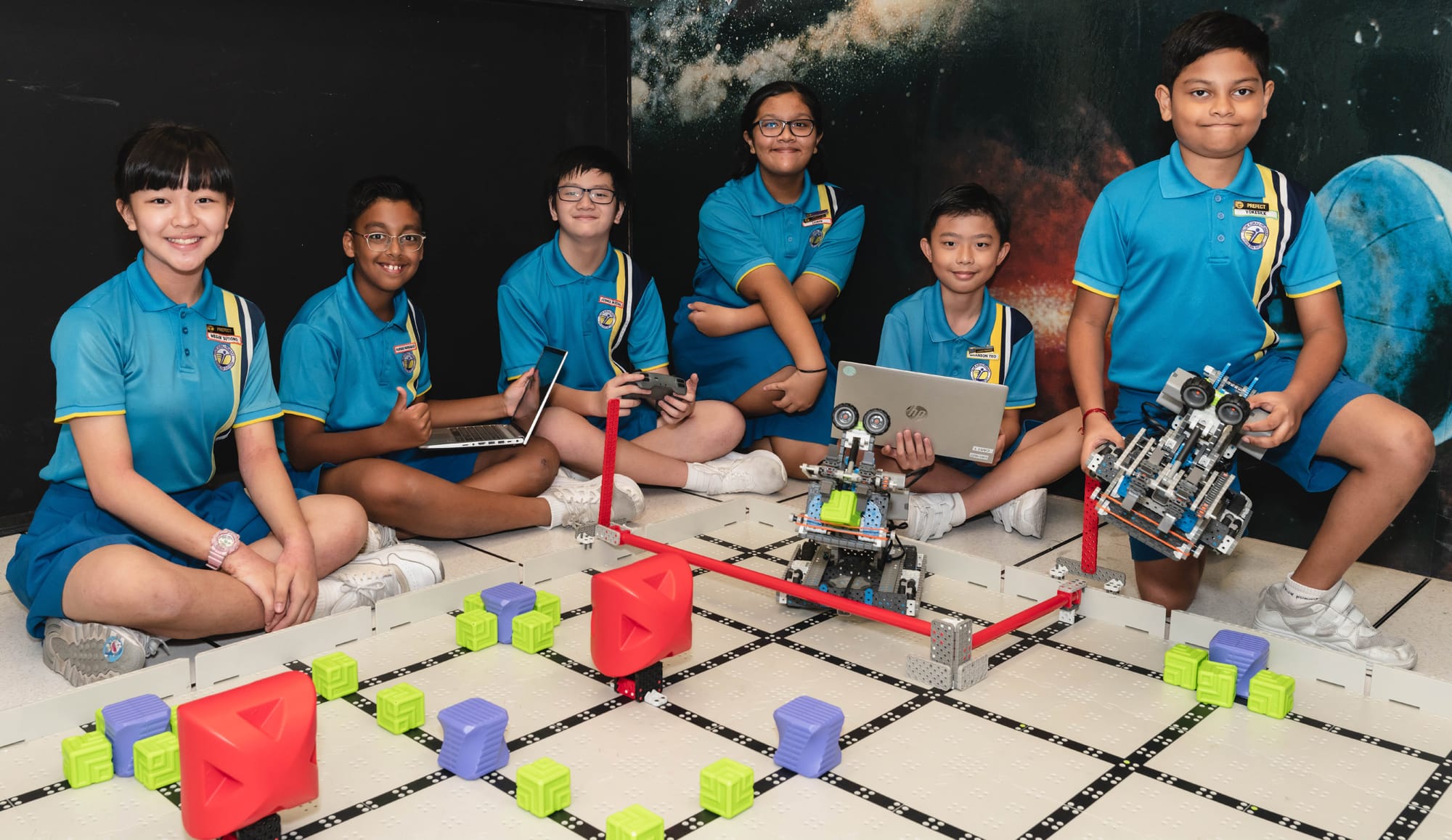 Six children in blue uniforms sit with laptops, built robots, and colorful blocks on a robotics competition mat.