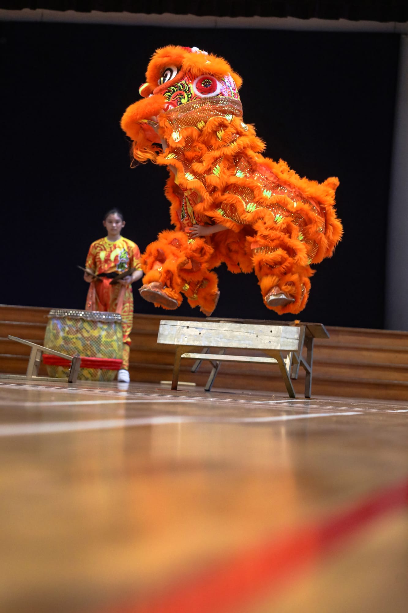 A vibrant orange Chinese lion dance costume leaps over a bench on a wooden stage, with a performer watching nearby.