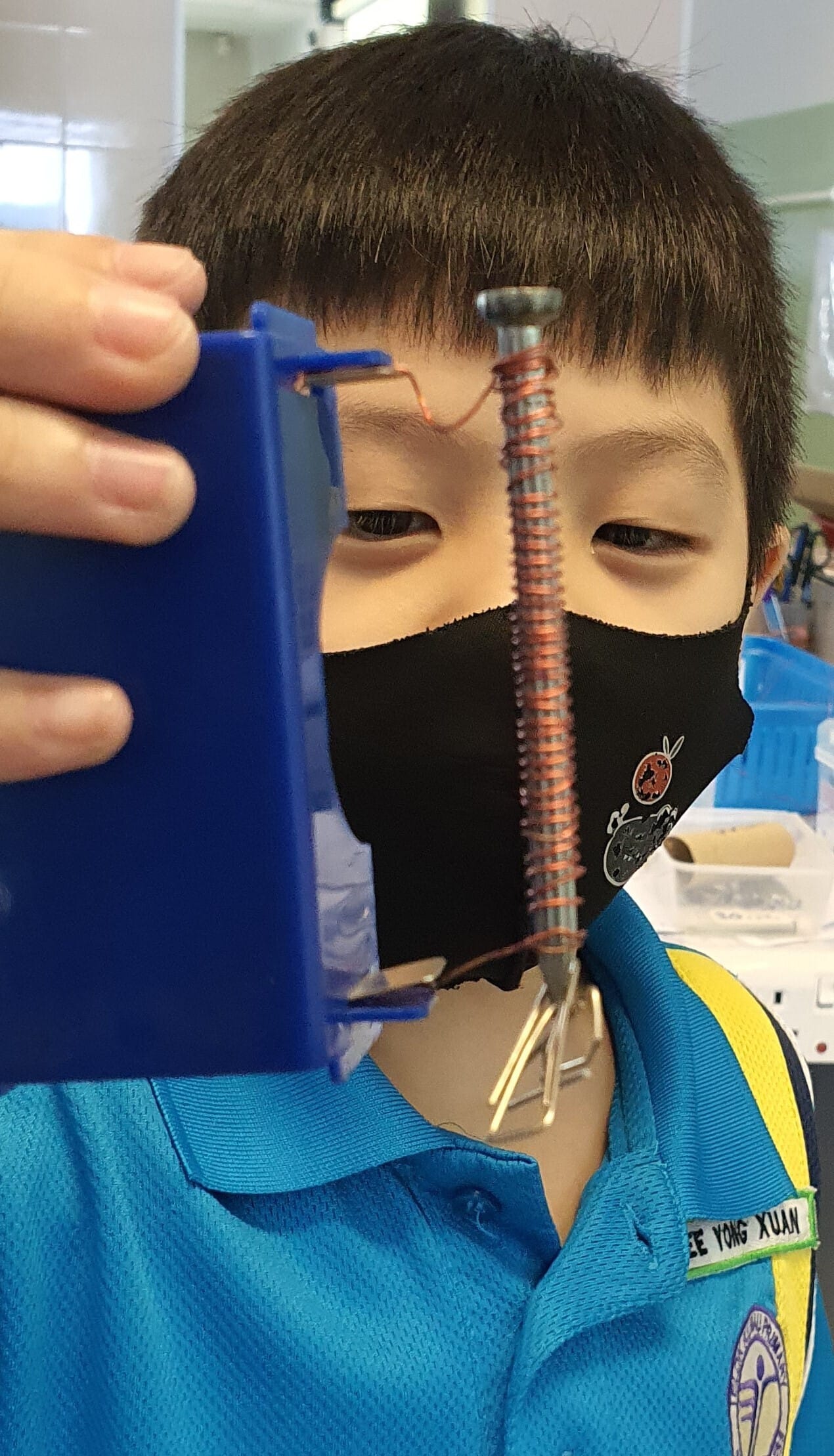A masked boy in a blue school uniform holds up a screw wrapped with copper wire coiled around it.