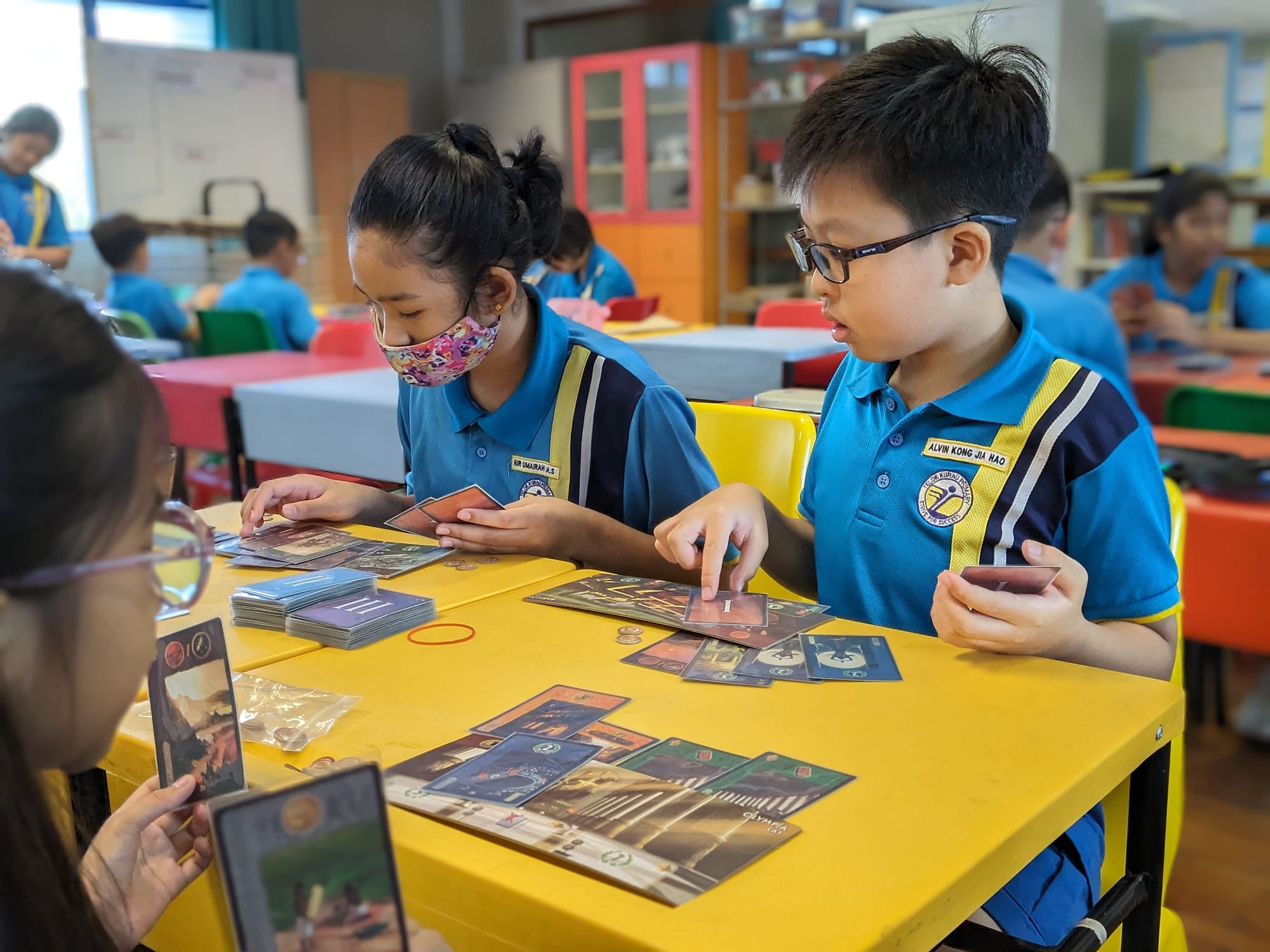 Three schoolchildren in blue uniforms play a card game at a yellow table in a classroom.