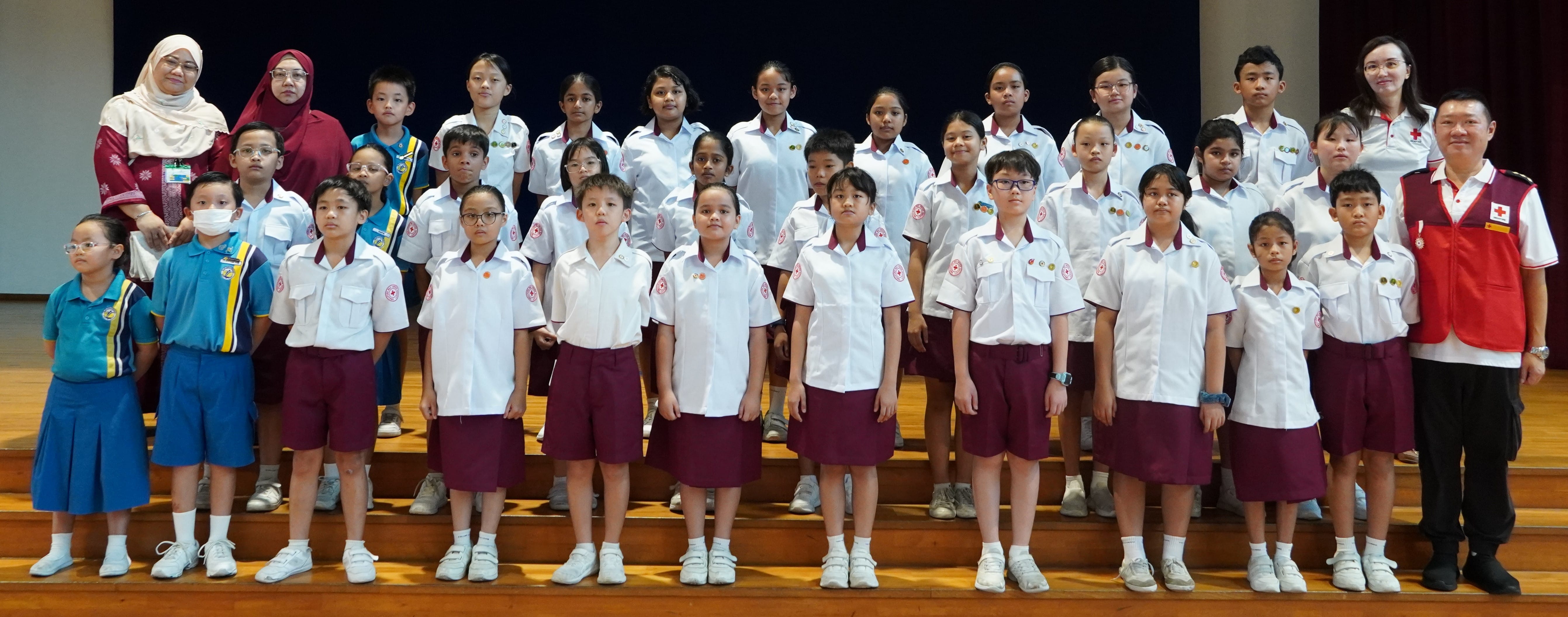 Group of school children in white and maroon uniforms posing on wooden steps with two Red Cross adult volunteers.