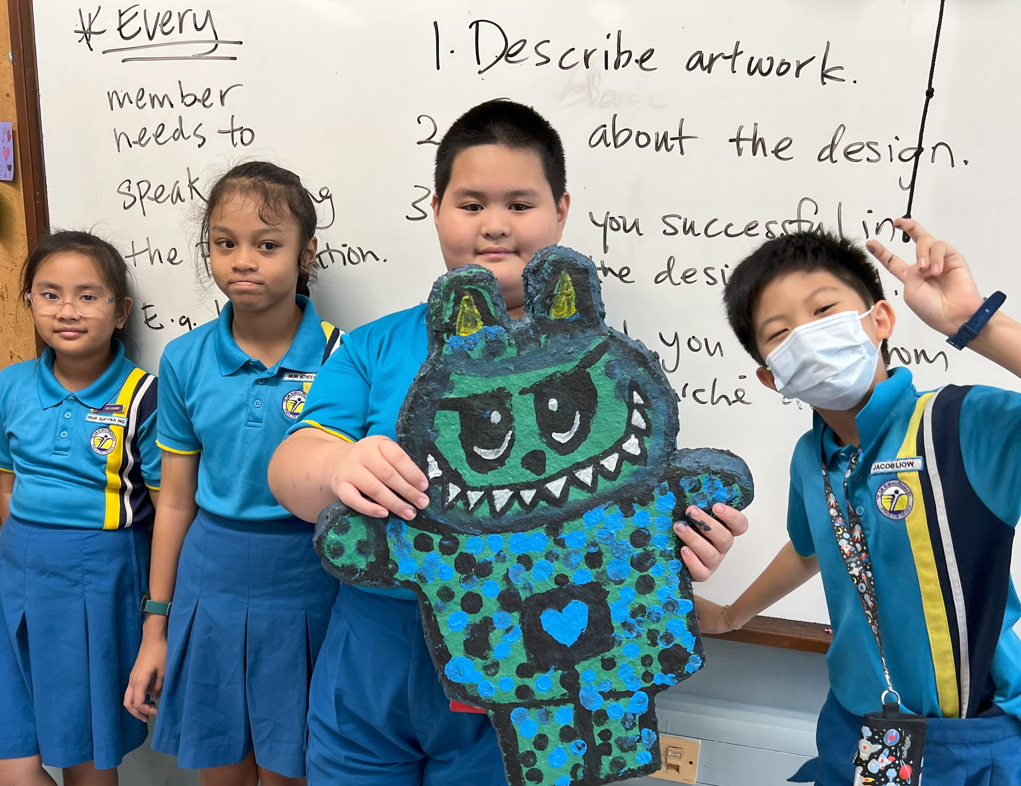 Four school children in blue uniforms stand before a whiteboard, holding a painted green and blue monster artwork.