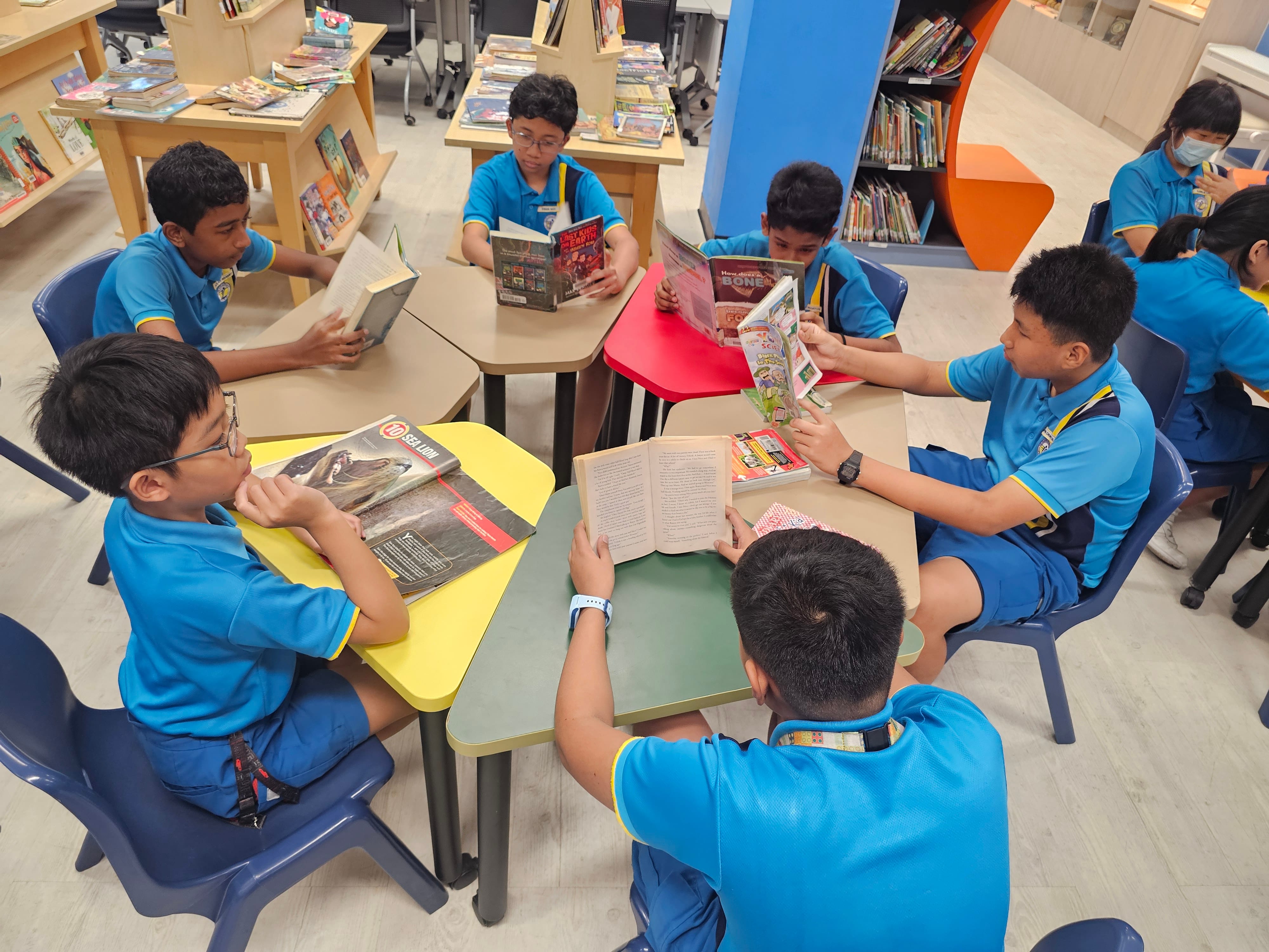 Several children in blue uniforms sit at colorful tables in a school library, each reading a different book.