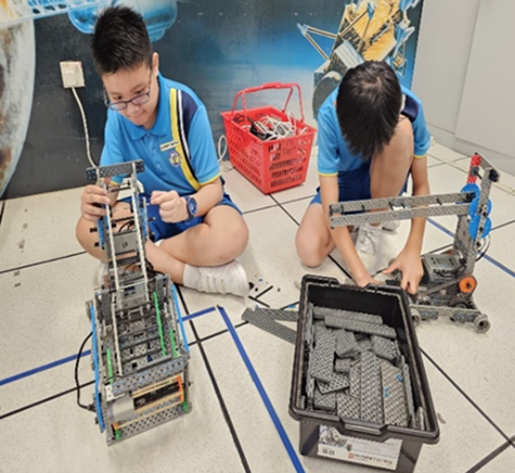 Two children in blue uniforms sit on a tiled floor assembling LEGO robots with parts in nearby containers.