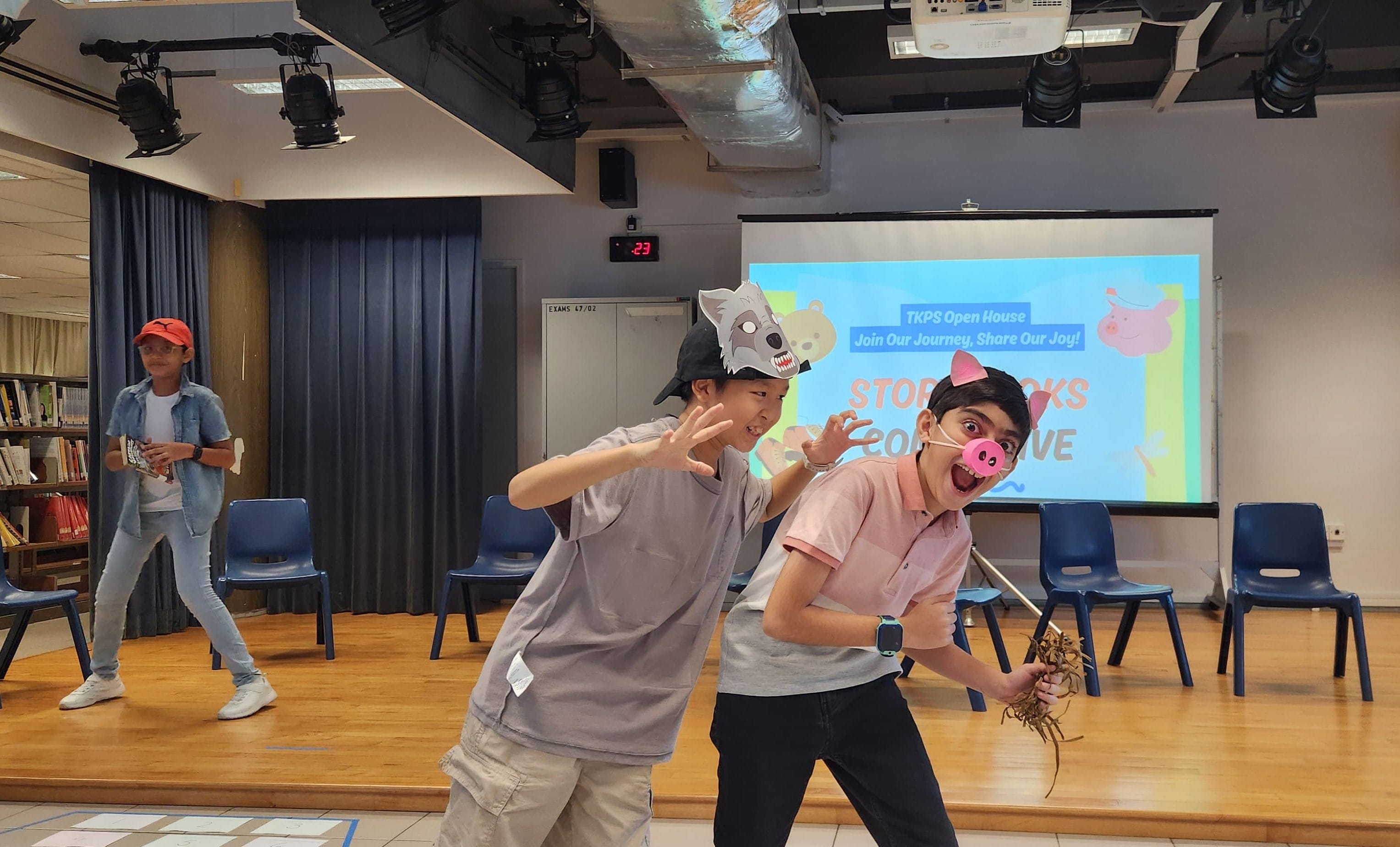 Three kids perform a Three Little Pigs skit; one wears a wolf mask, another a pig nose, in a school hall.
