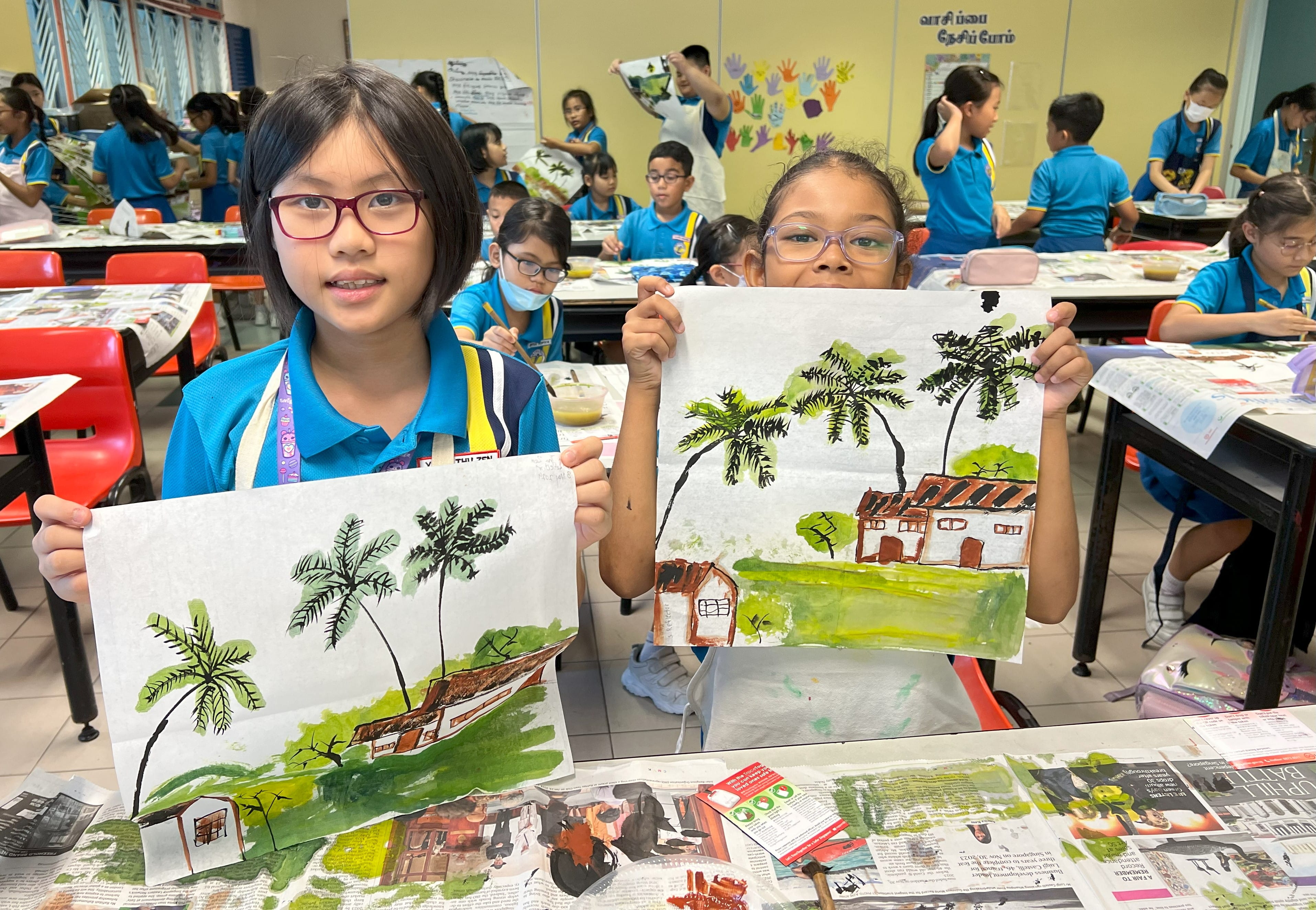 Two schoolgirls in blue uniforms hold up watercolor paintings of houses with palm trees during an art class.