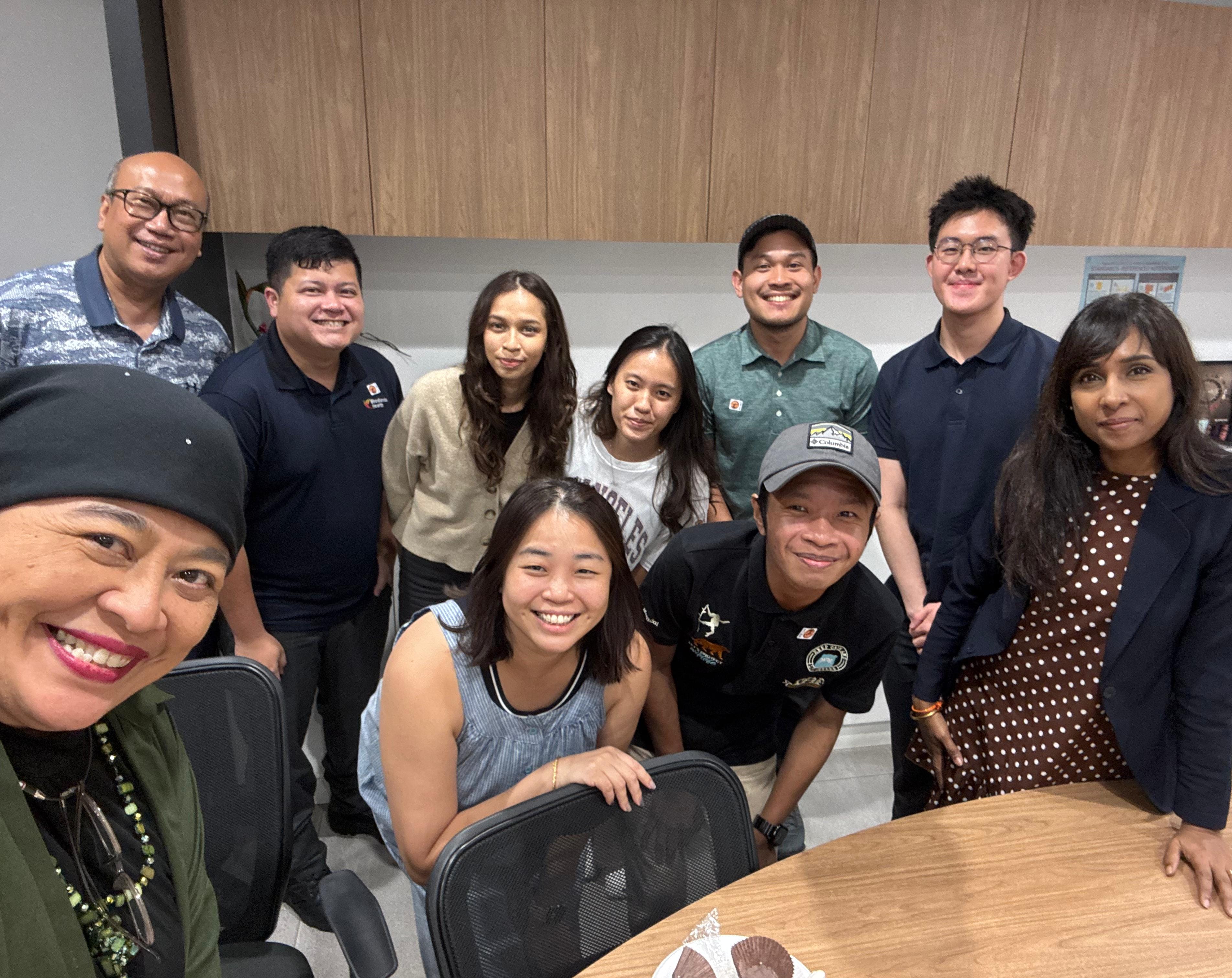 Ten people smiling together in an office room around a wooden table, posing for a group selfie.