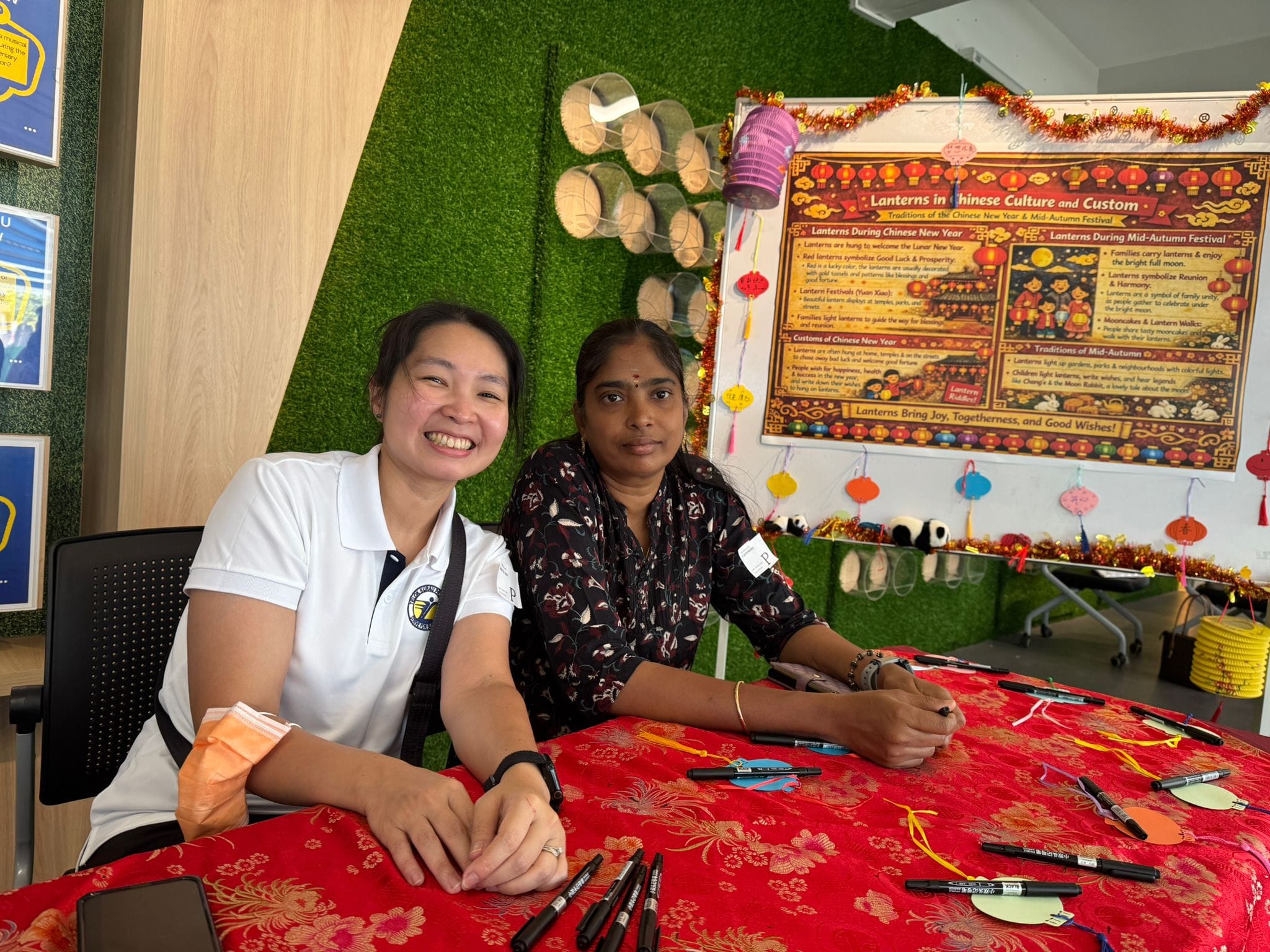 Two women sit at a red table with markers at a Chinese lantern festival cultural display booth.