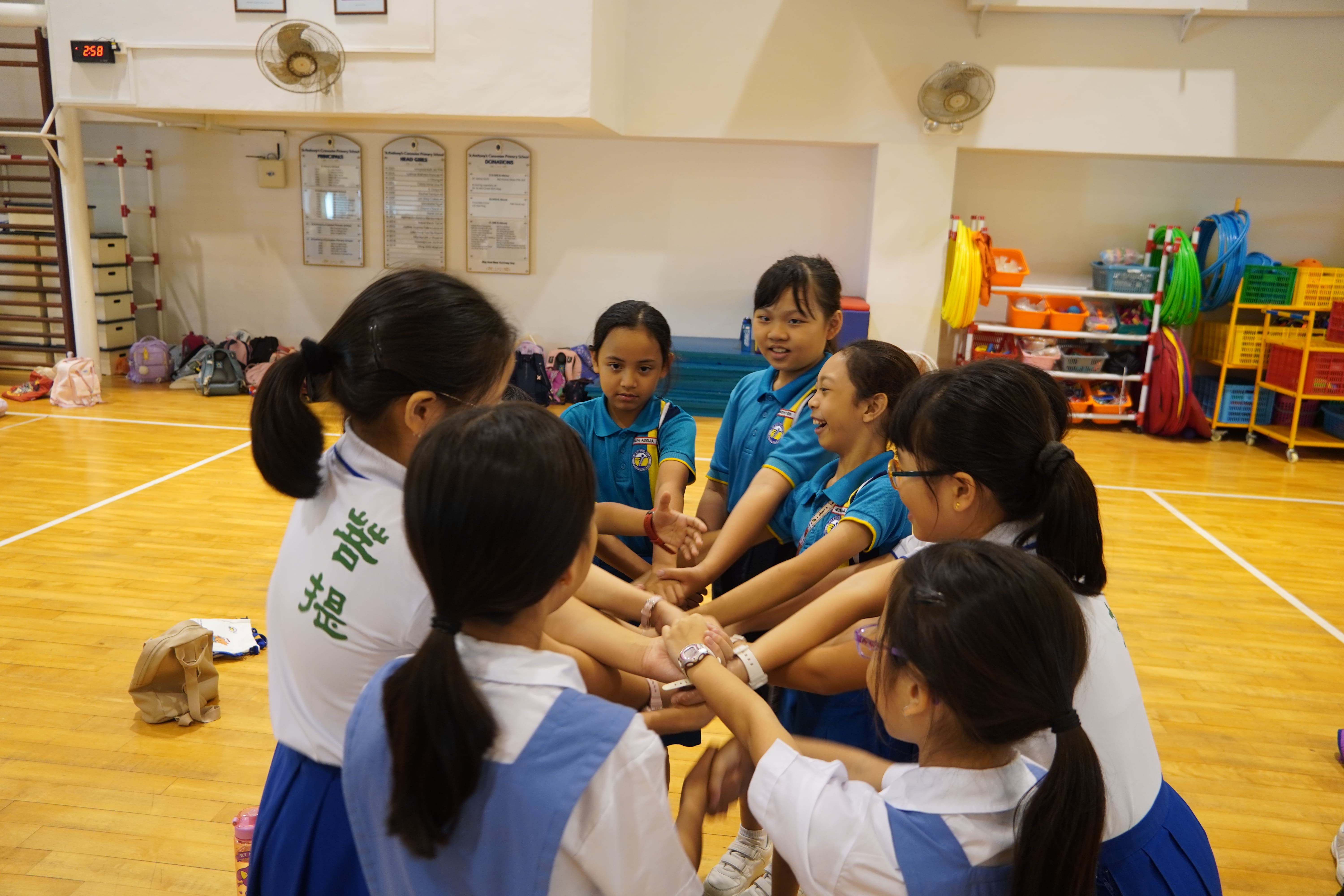 A group of young schoolgirls in uniforms sitting in a circle on a gym floor, stacking hands together.