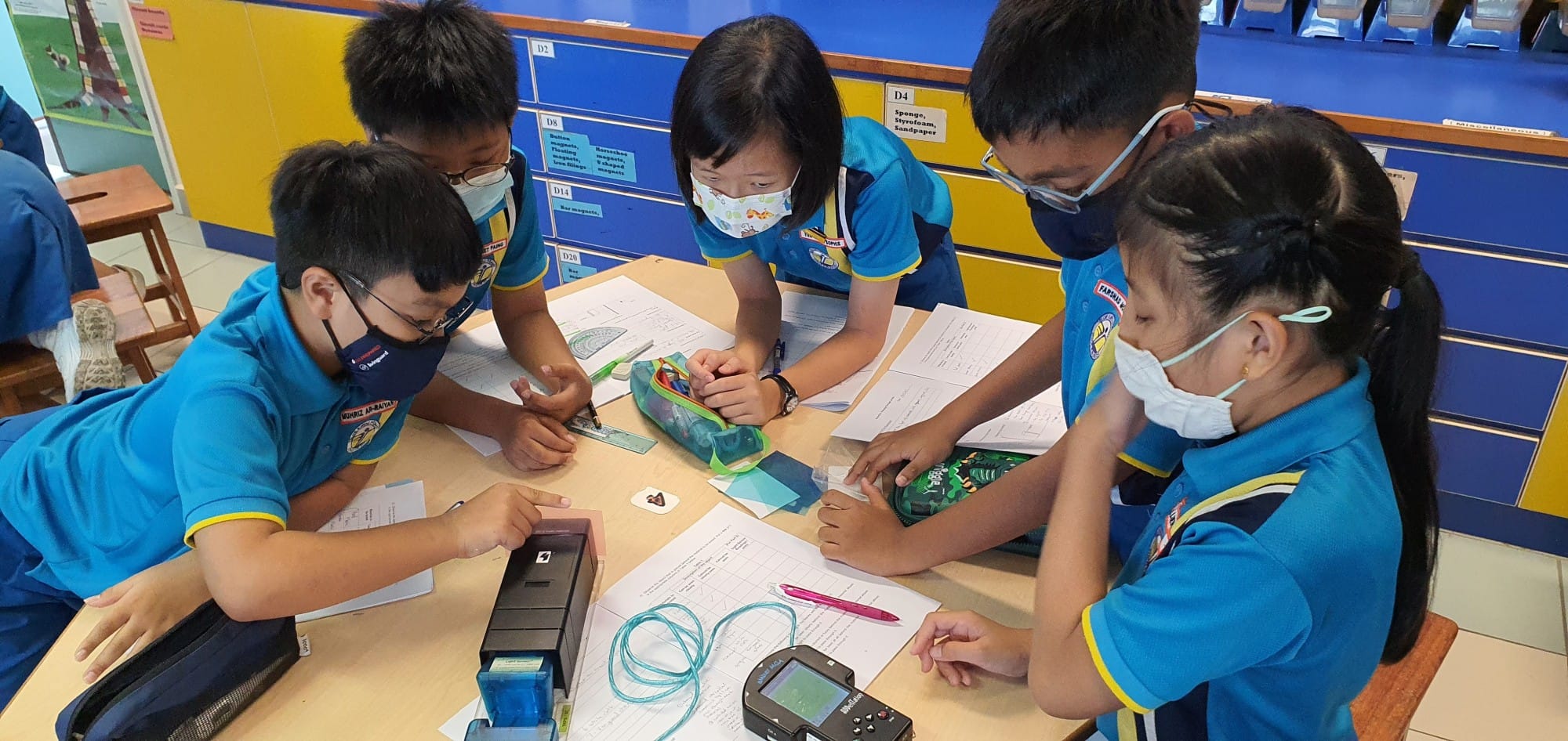 Five masked children in blue uniforms gather around a desk doing a science activity with worksheets and equipment.