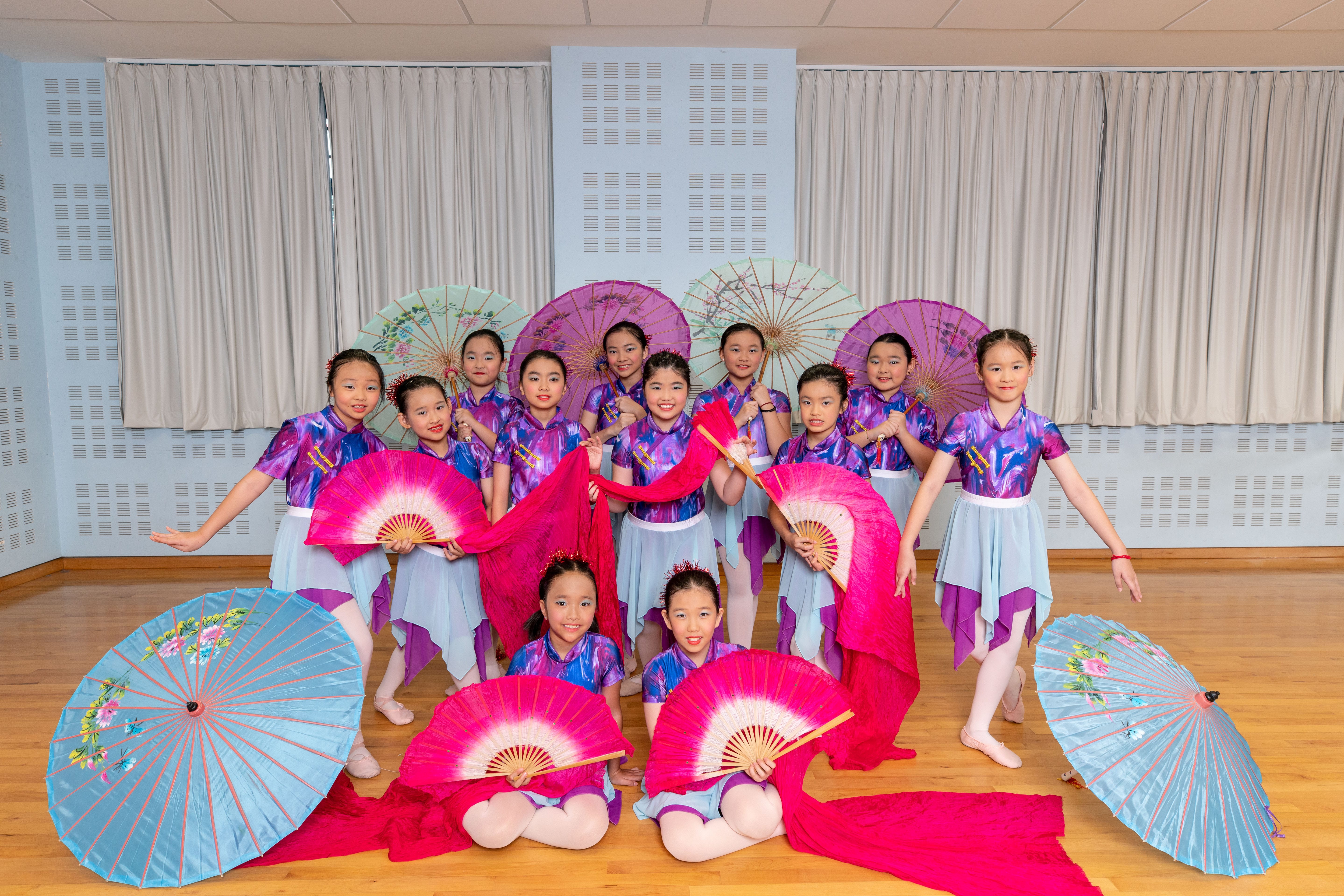 Young girls in purple costumes pose with large pink fans and colorful parasols in a dance studio.