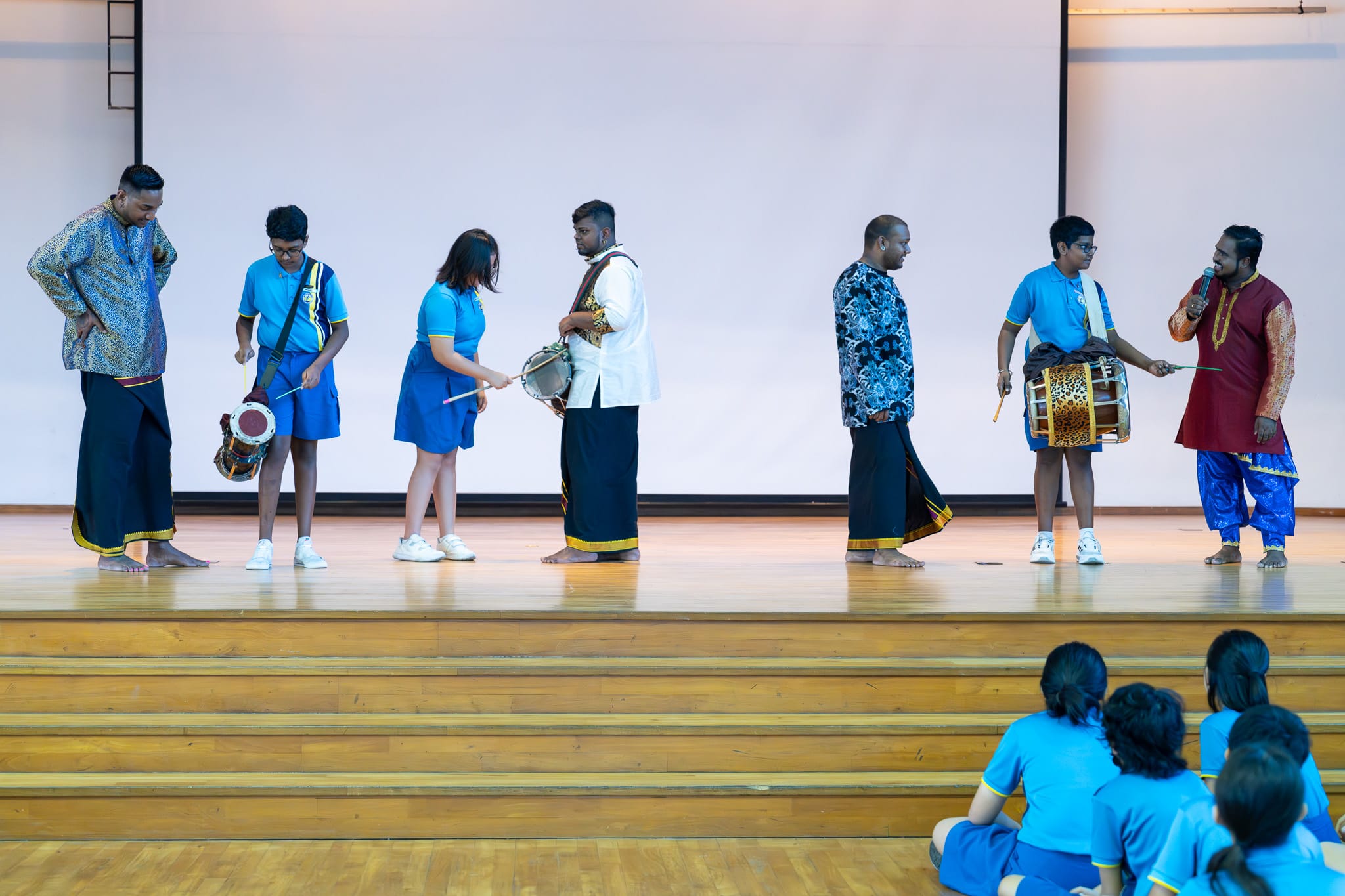 Seven people on a wooden stage learn to play traditional drums, watched by seated students in blue uniforms.