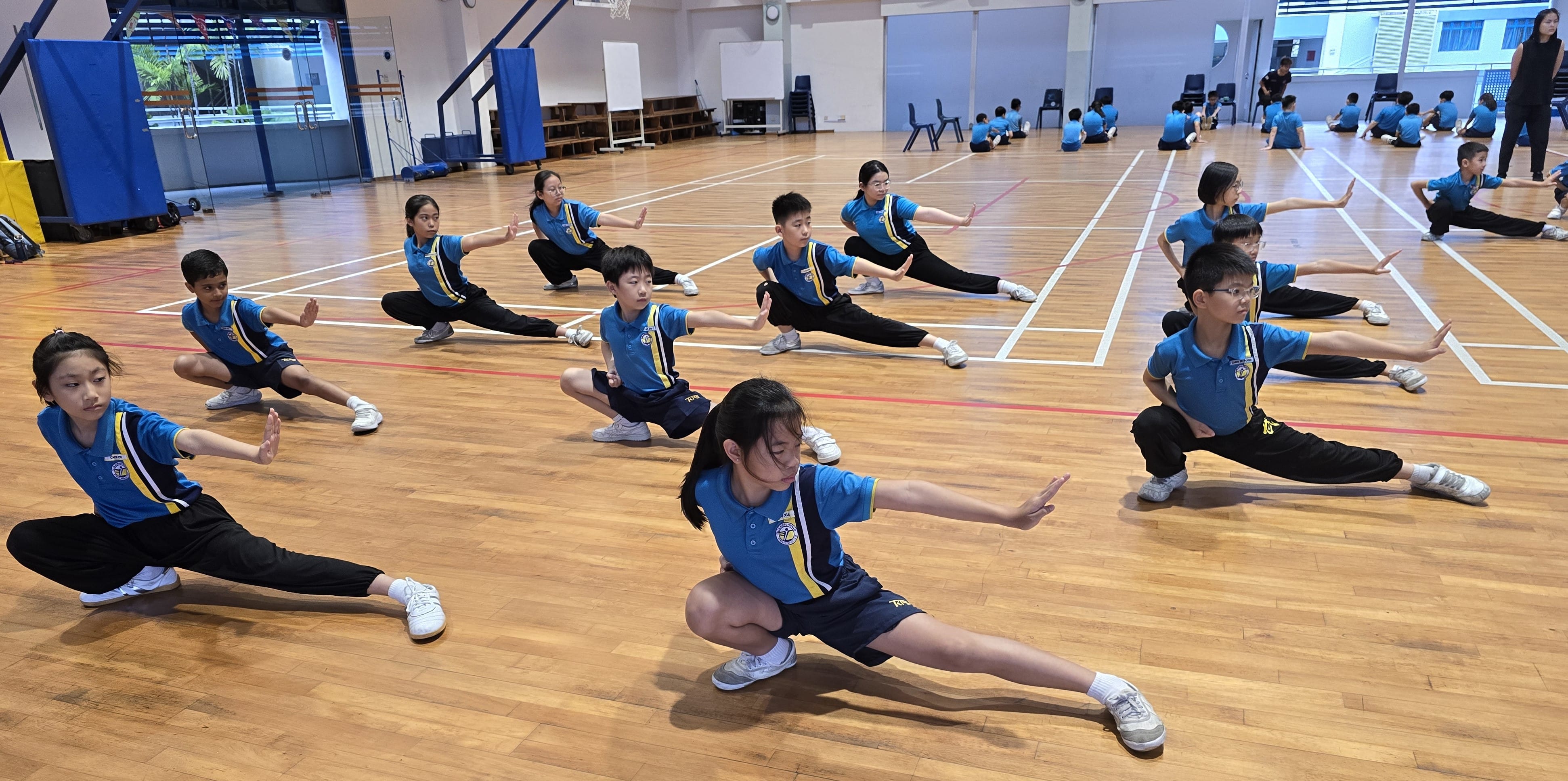 Students in blue uniforms practice wushu stances in a gymnasium, arms extended and legs in wide lunges.