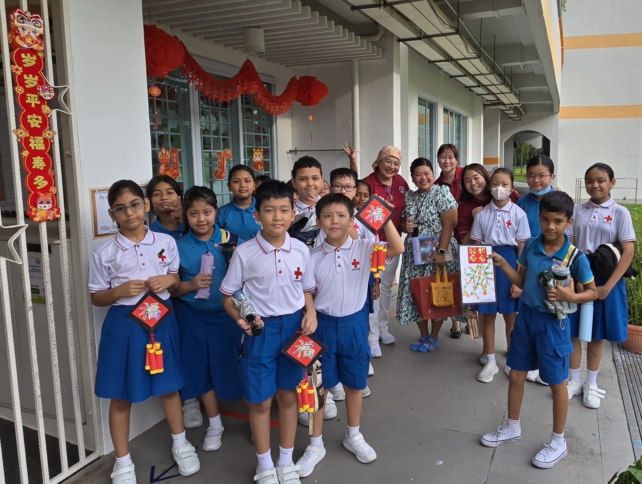 A group of schoolchildren in uniforms and adults pose outdoors near Chinese New Year decorations, holding crafts and cards.