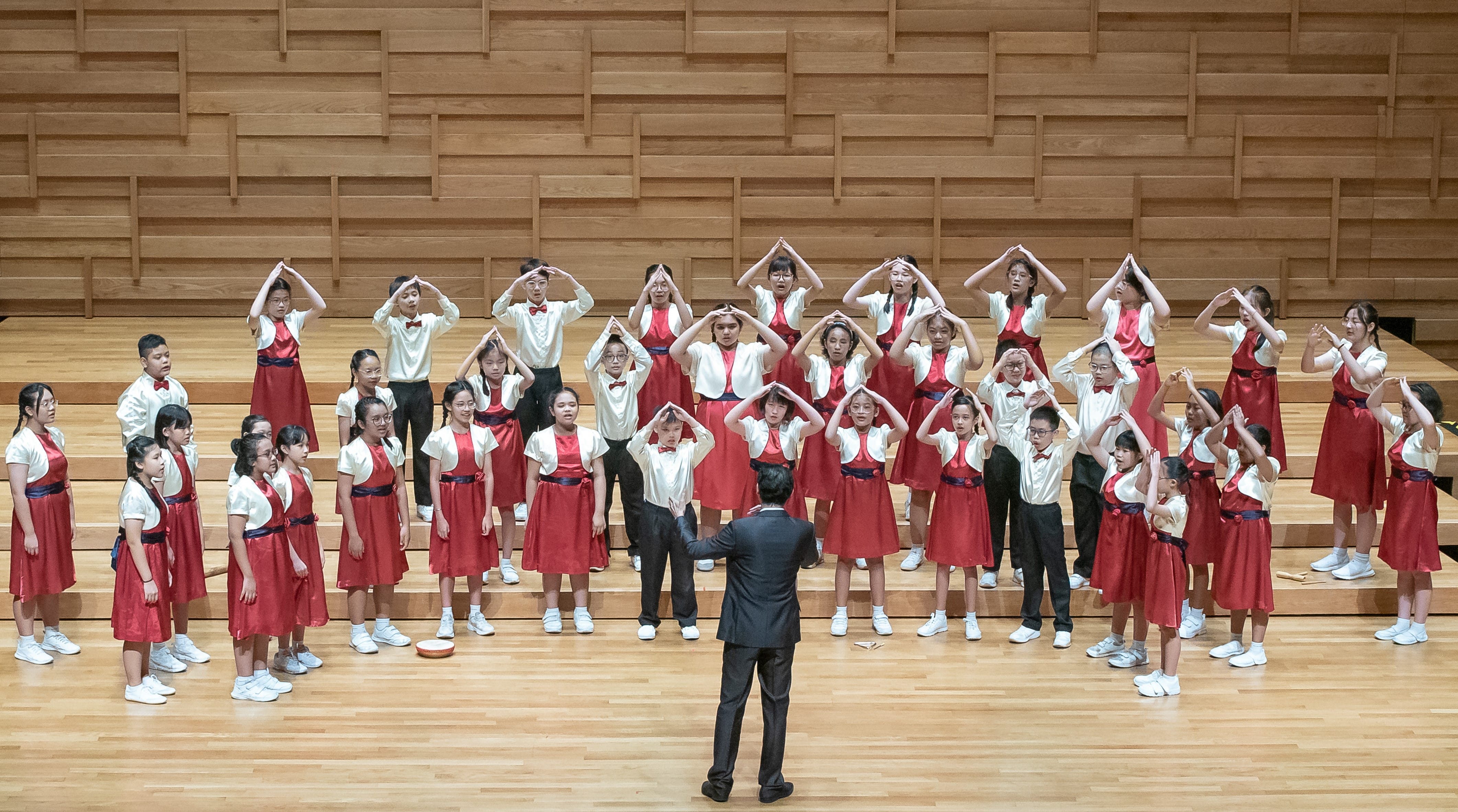 A children's choir in red and white uniforms performs on a wooden stage, arms raised, led by a conductor in a dark suit.