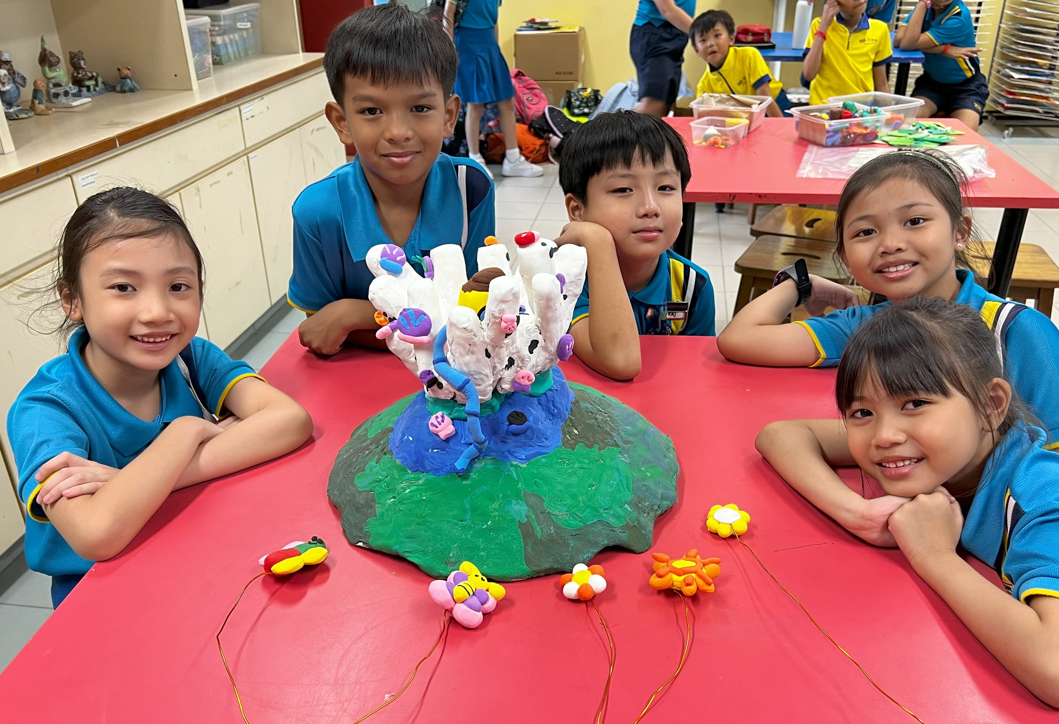 Four children in blue uniforms smile around a red table displaying a colorful clay coral reef sculpture.