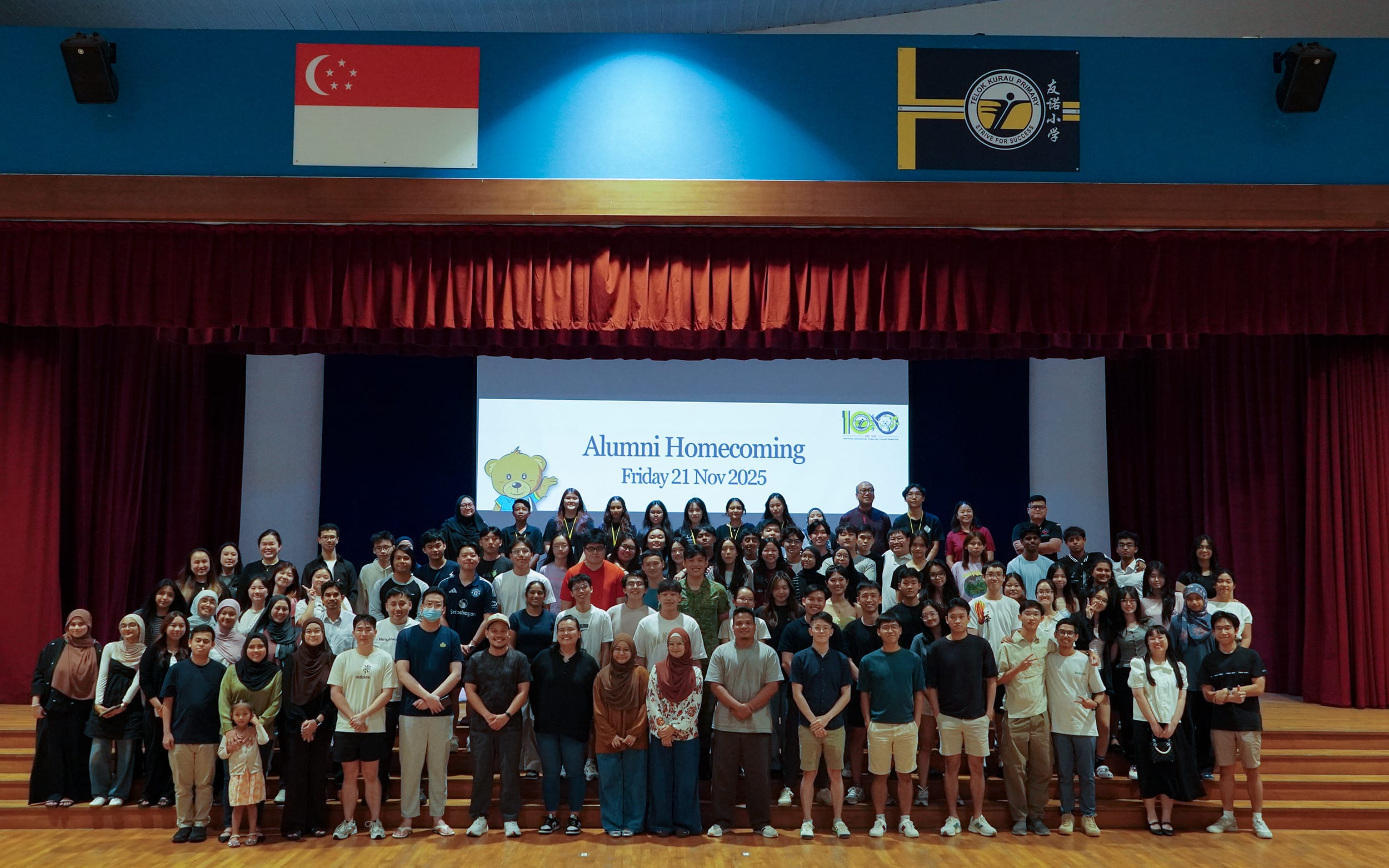 Large group posing in a hall before a screen reading Alumni Homecoming Friday 21 Nov 2025, with Singapore flag above.