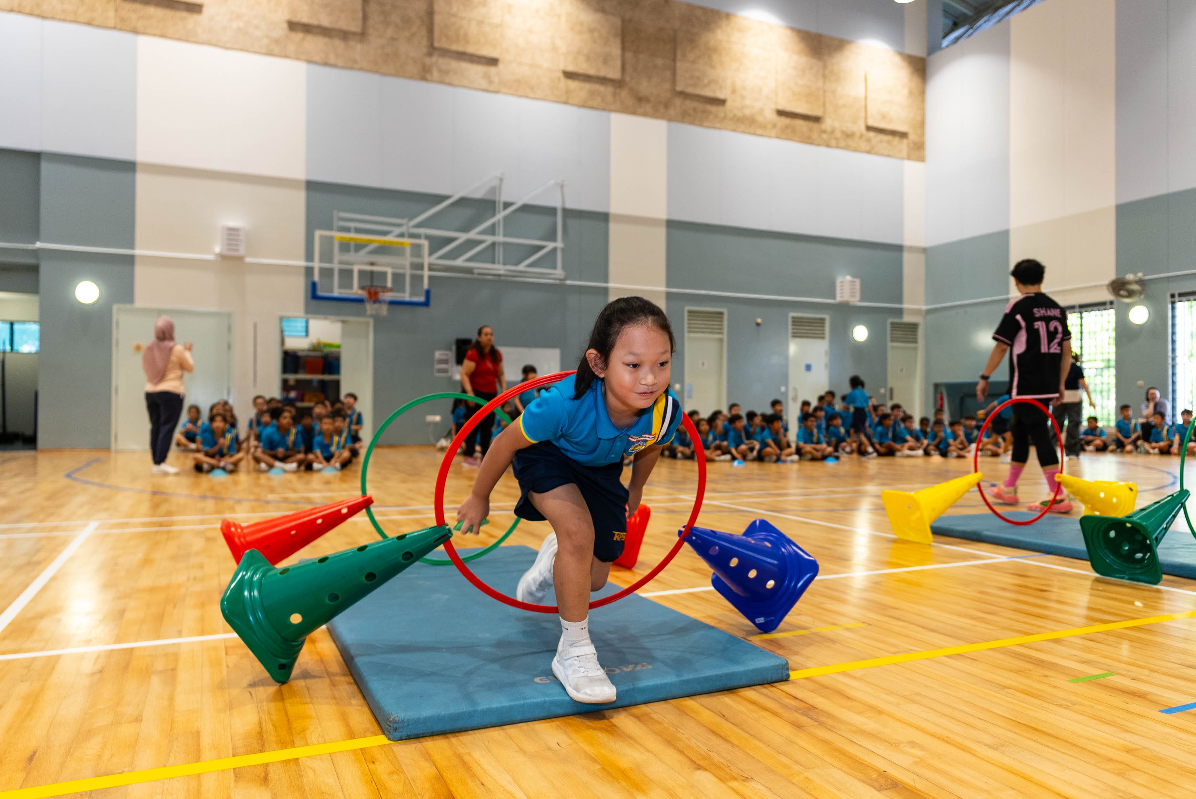 A young girl in a blue uniform crawls through a hoop on an obstacle course in a school gymnasium.