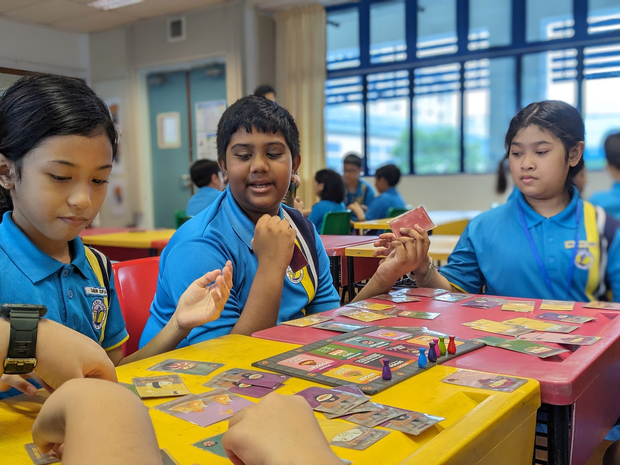 Three children in blue school uniforms playing a board game with cards and colored pawns on a yellow table.