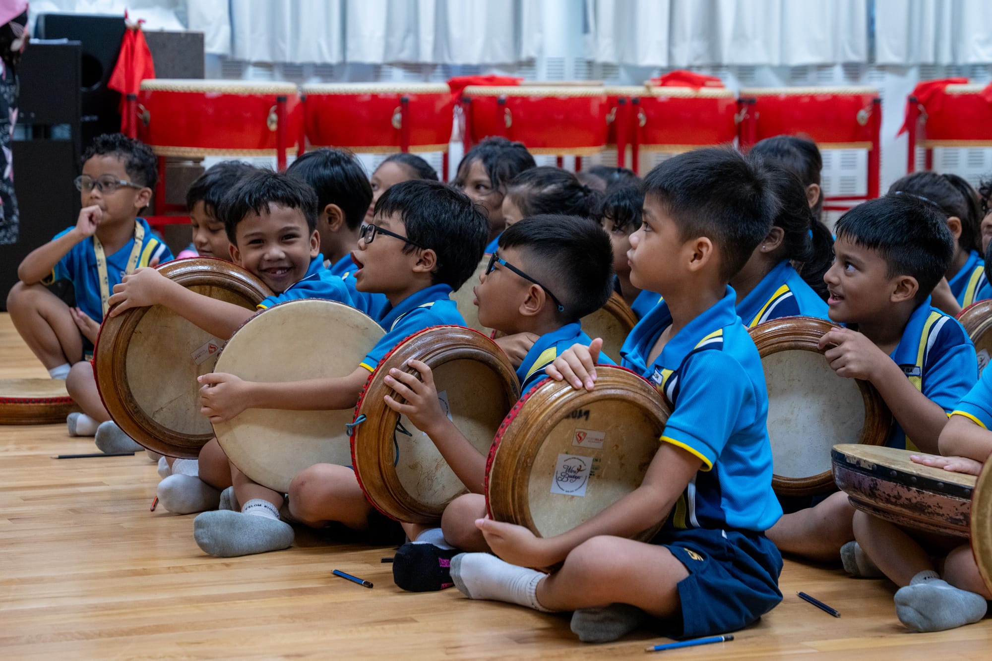 Young children in blue uniforms sit cross-legged on a wooden floor, each holding a small round drum.