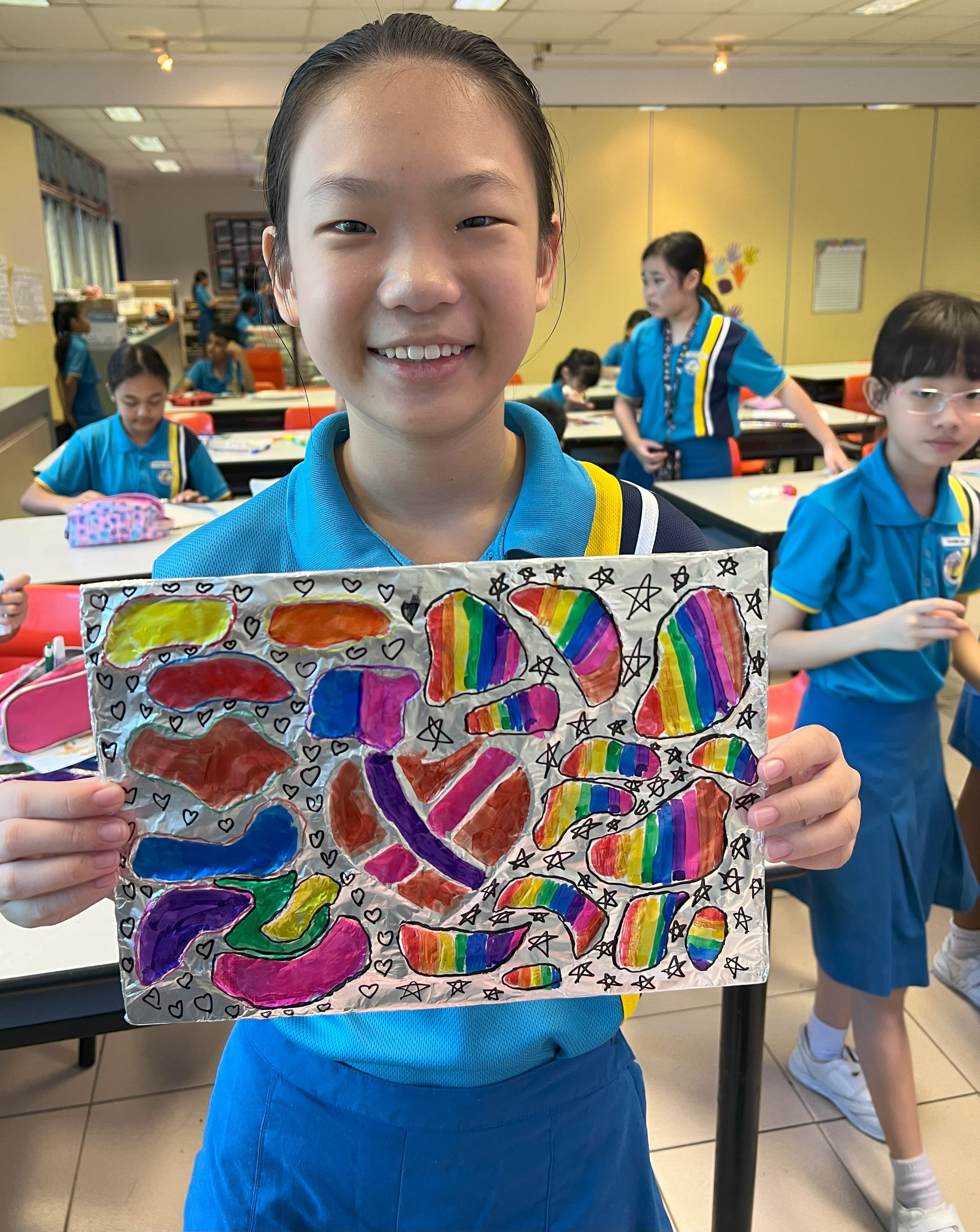 A smiling girl in a blue school uniform holds up a colorful foil artwork with rainbow shapes, hearts, and stars.