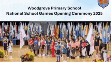 Children standing in a gym before many flags under a banner for Woodgrove Primary School 2025 opening ceremony