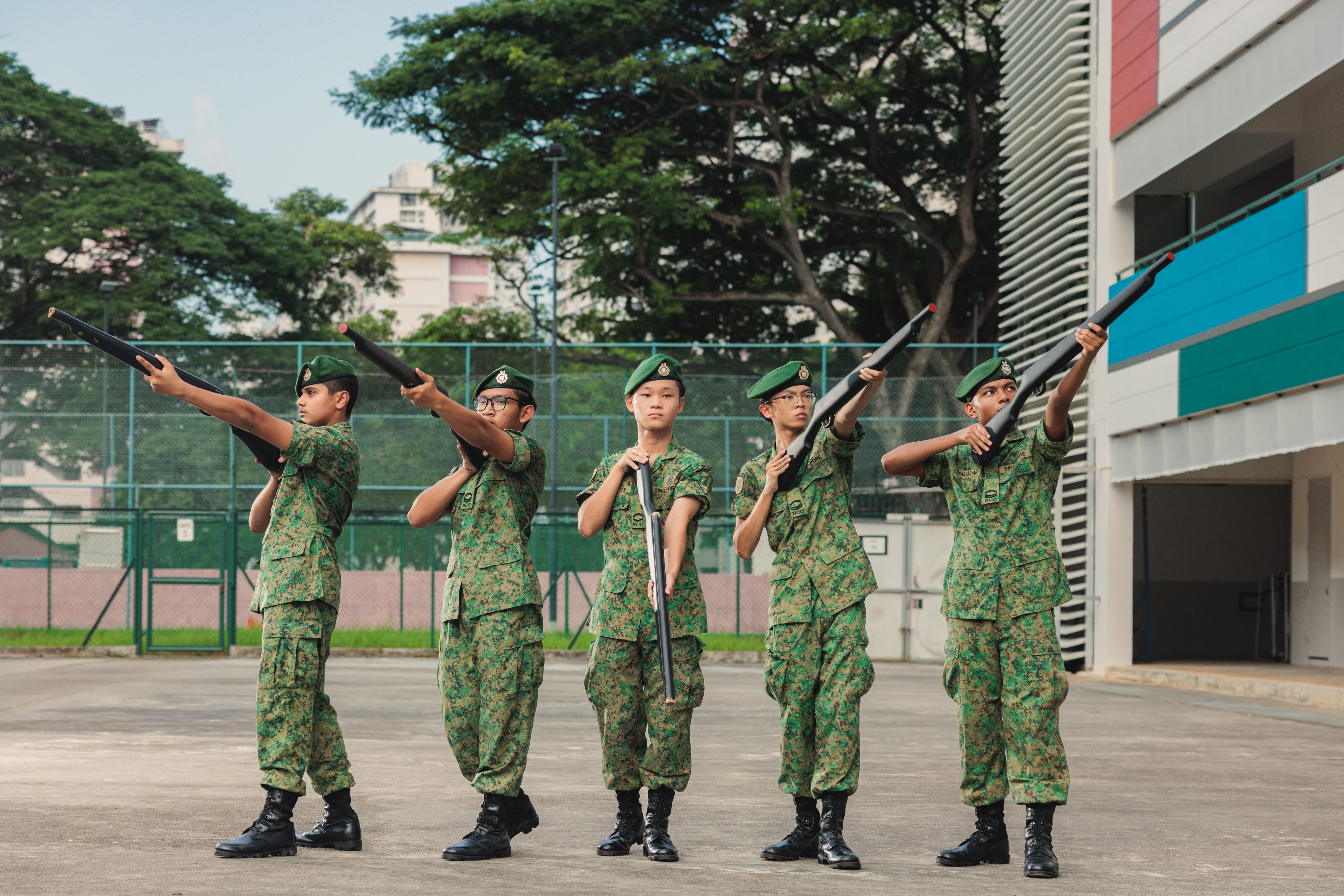 Five young cadets in green camouflage uniforms and berets hold rifles raised during an outdoor drill.