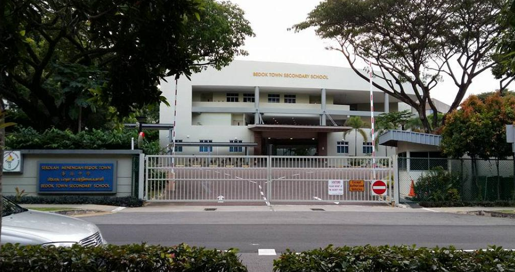 Front entrance of Bedok Town Secondary School with closed metal gates, a sign, and surrounding trees.