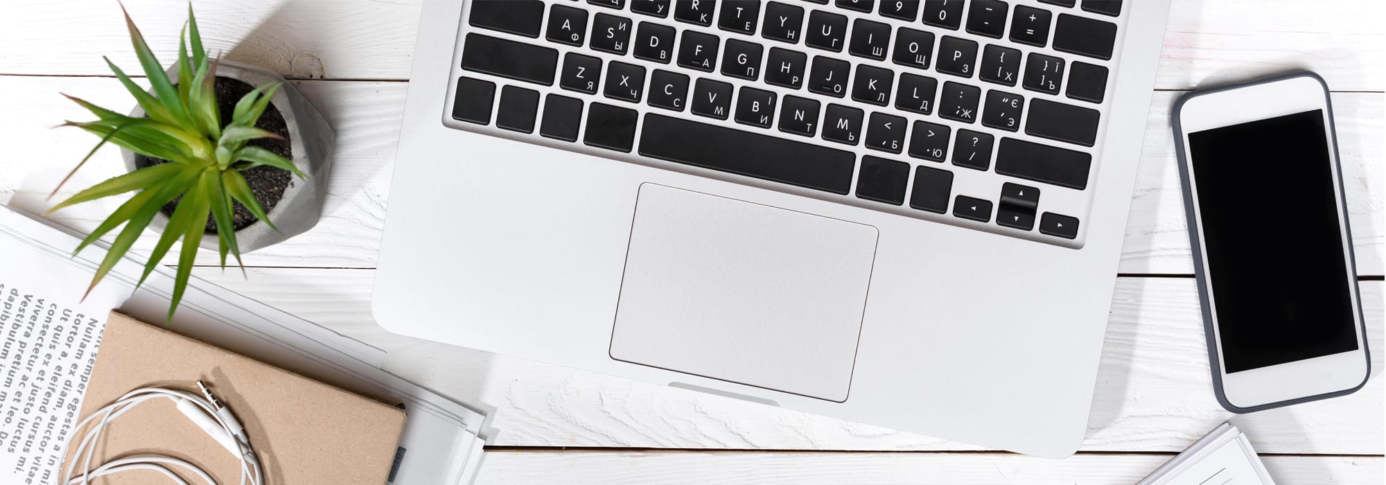 Overhead view of a laptop, smartphone, small plant, notebook, and earphones on a white wooden desk.