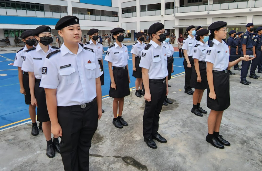 Young cadets in white shirts, black berets, and masks stand in formation on an outdoor school parade square.