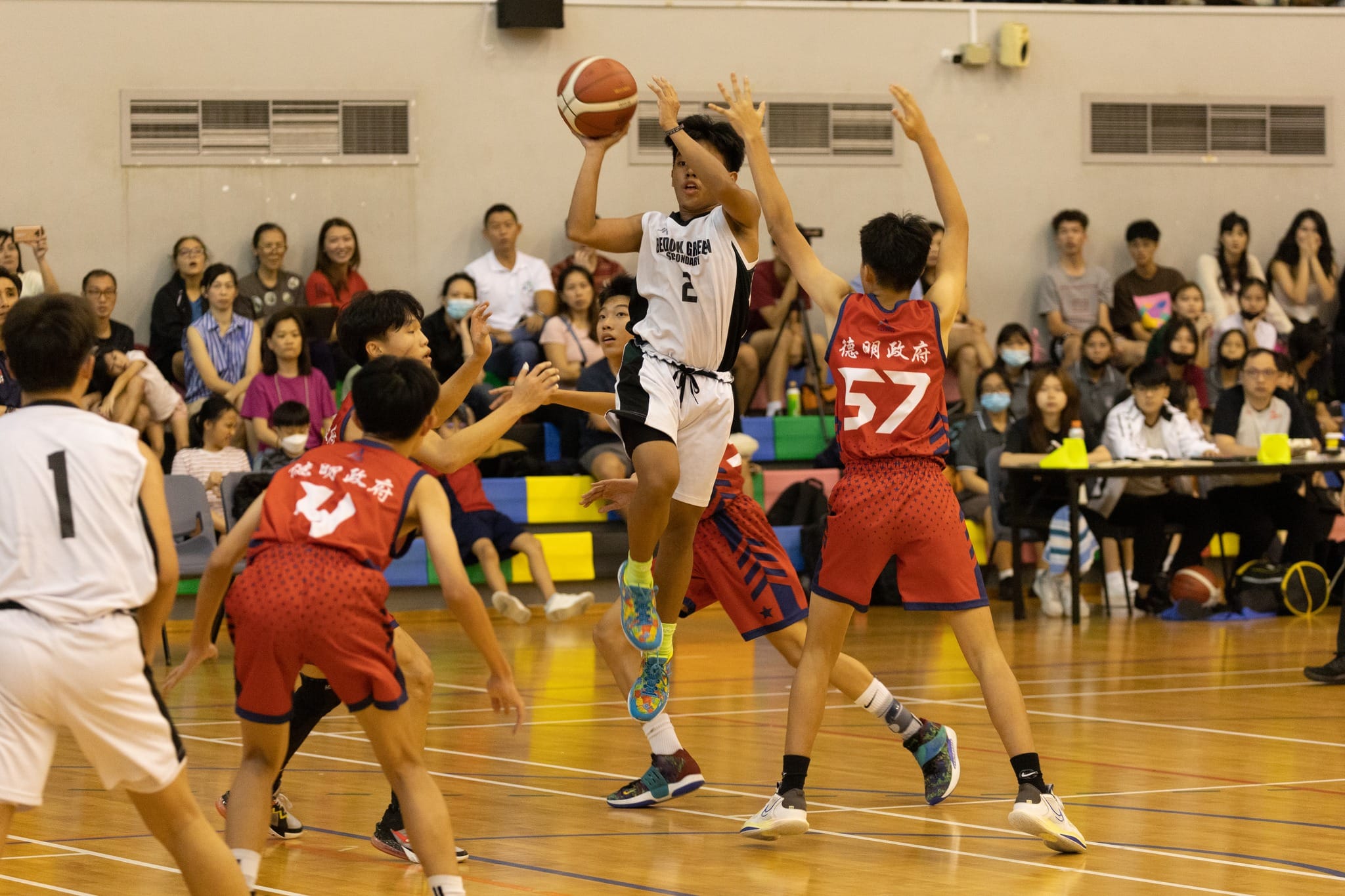 A youth basketball player in white jersey number 2 jumps to shoot while defenders in red uniforms contest the shot.