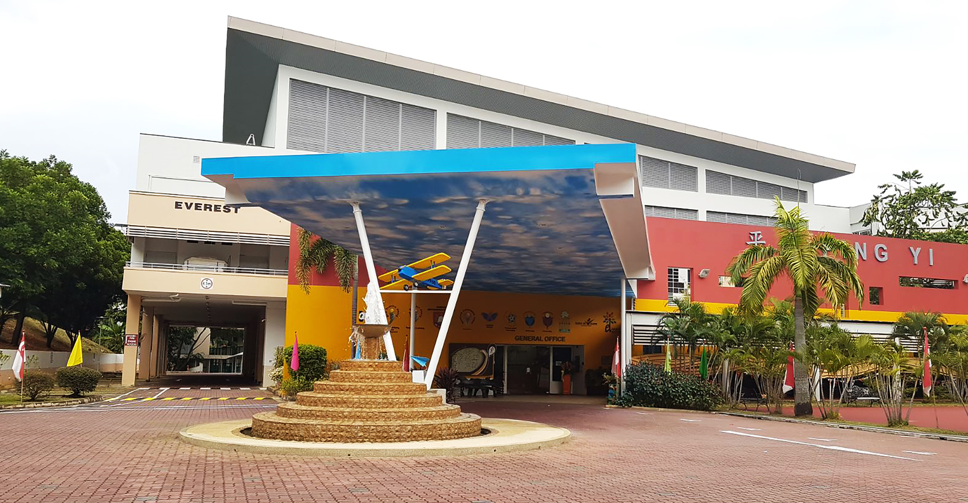 School entrance with a blue canopy, circular fountain, yellow walls, biplane decoration, and signs reading Everest and General Office.