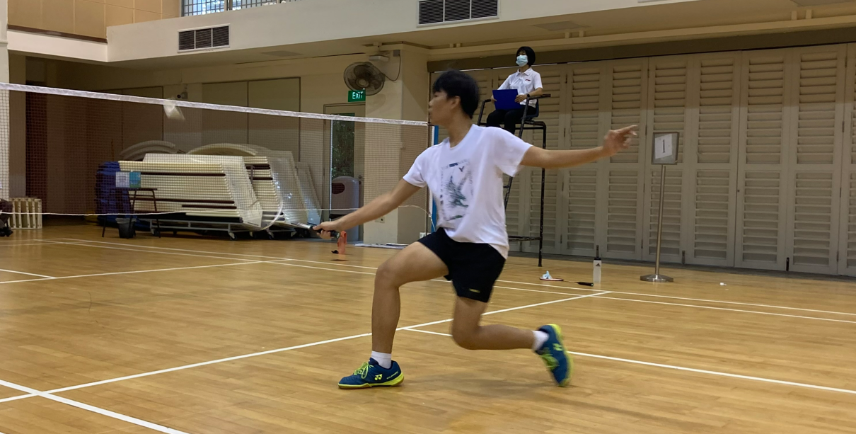 A young player lunges forward with a badminton racket near the net on an indoor wooden court, with a masked umpire seated behind.