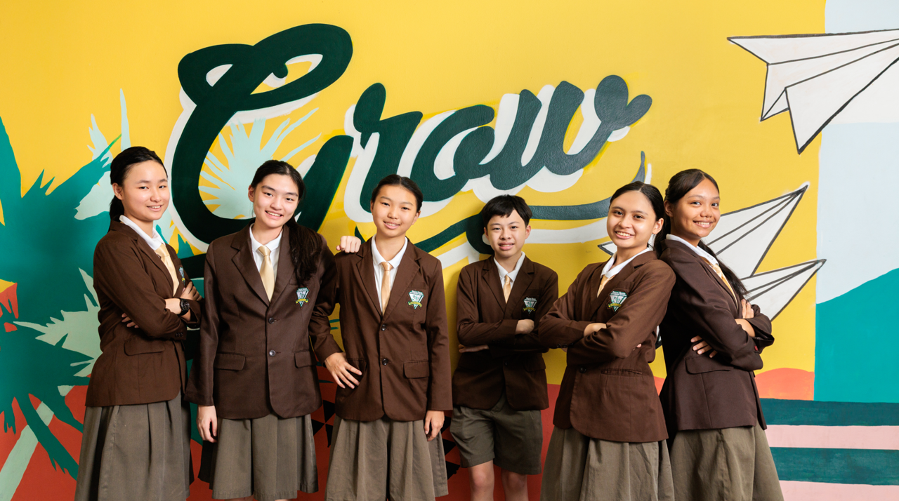 Six students in brown school uniforms smile in front of a colorful mural with the word Grow written on it.