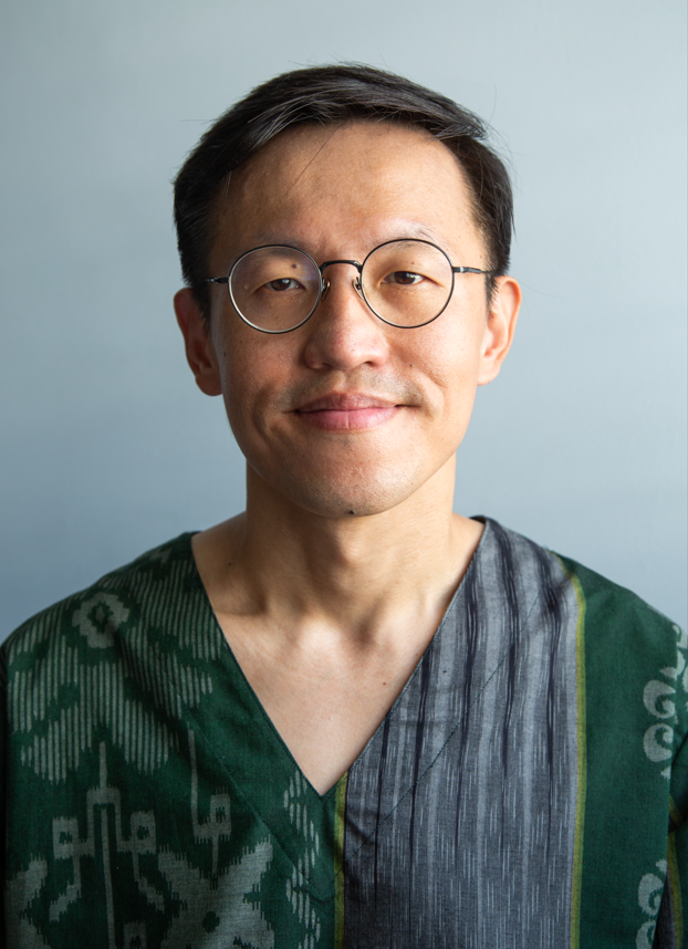 A man with round wire-rimmed glasses and short black hair smiling, wearing a patterned green and gray shirt.