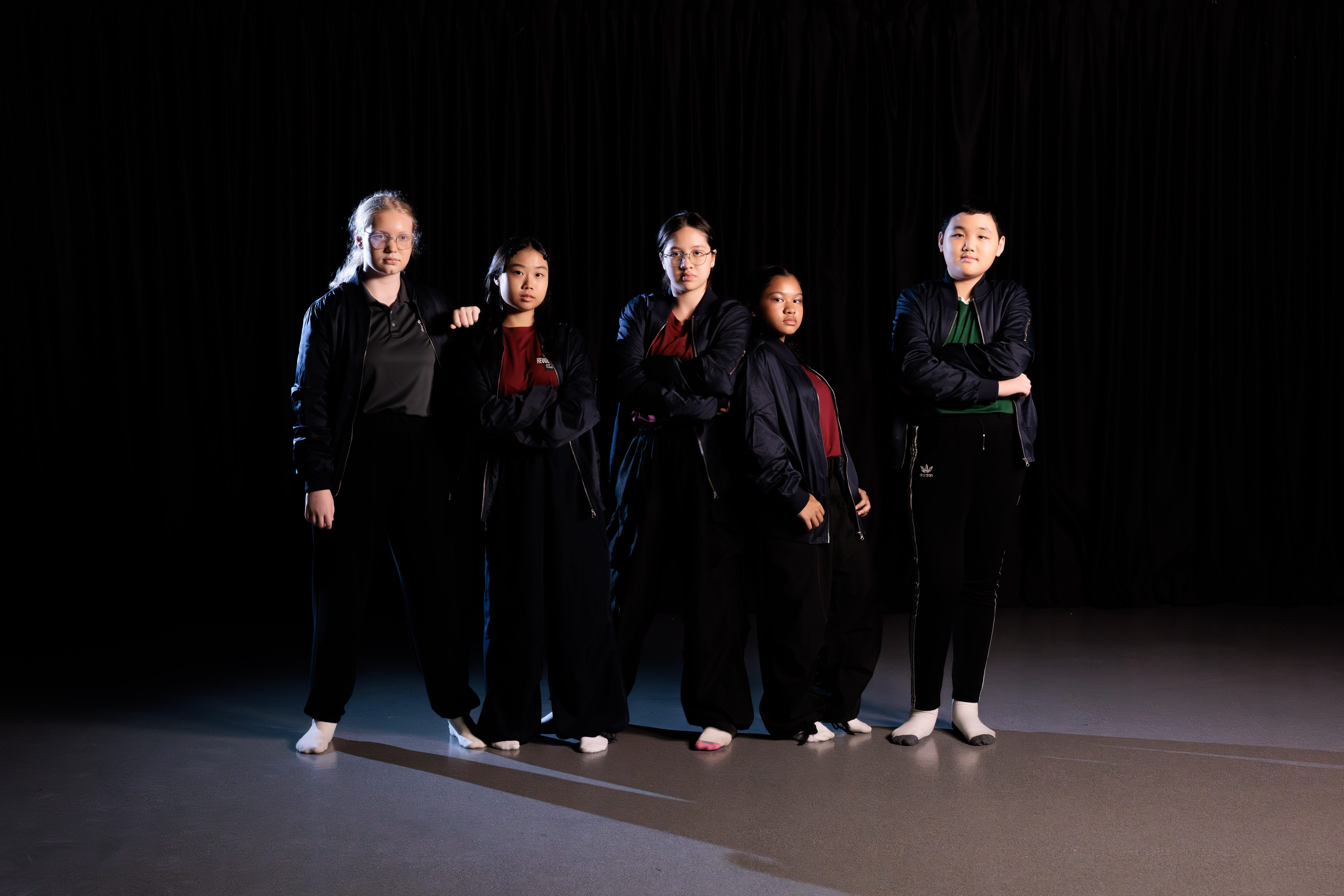 Five young people in black jackets and dark pants pose confidently against a black curtain backdrop under dramatic lighting.