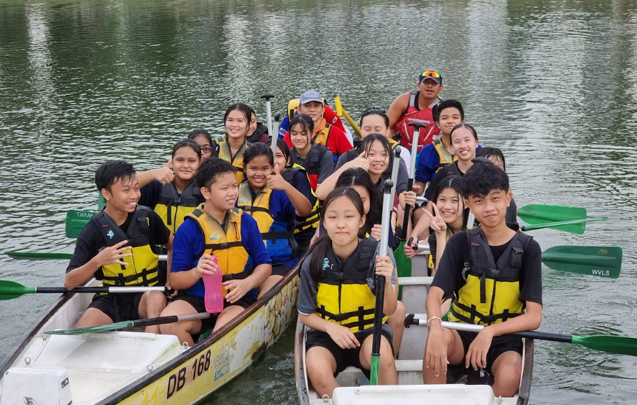 A group of young students wearing yellow life jackets sit in a dragon boat on calm water, holding paddles.