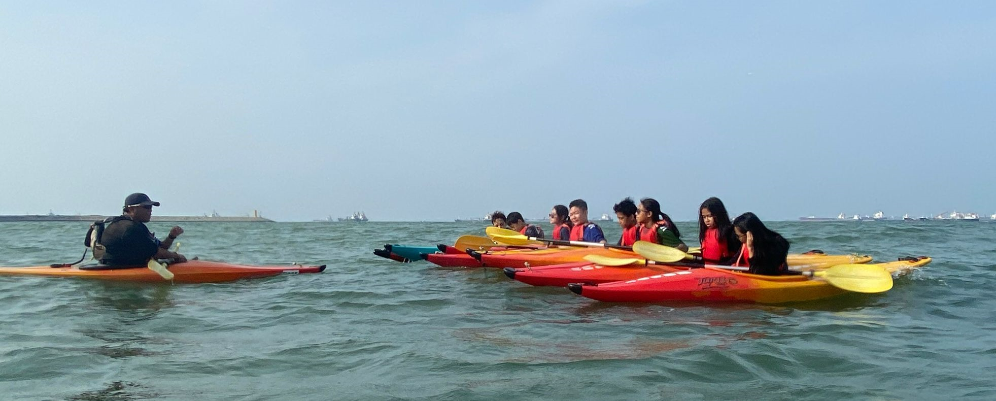 A group of young kayakers in red life jackets paddling colorful kayaks on open water, facing an instructor.