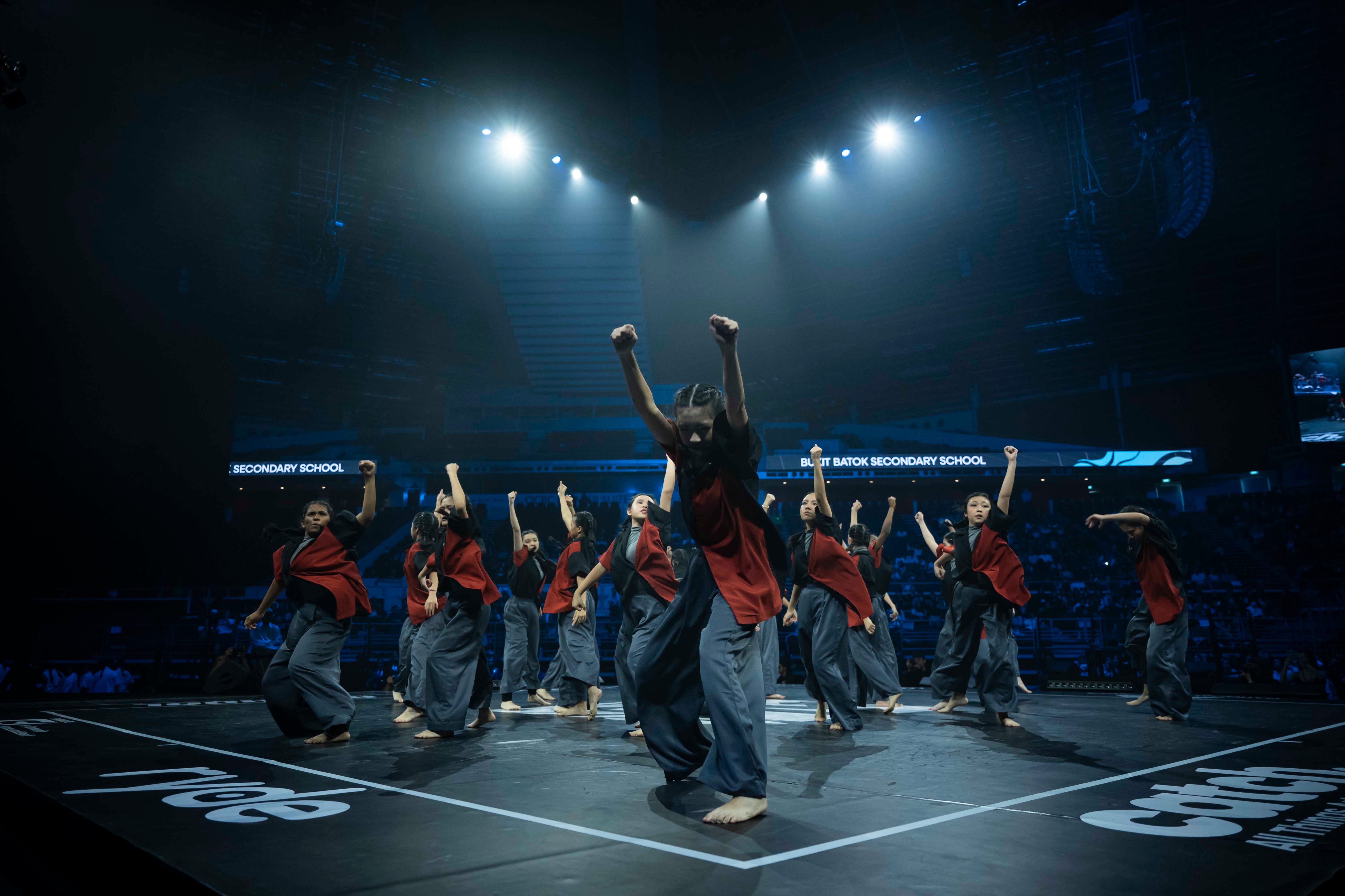A large group of dancers in red and black costumes perform on a dark stage under bright spotlights in an arena.