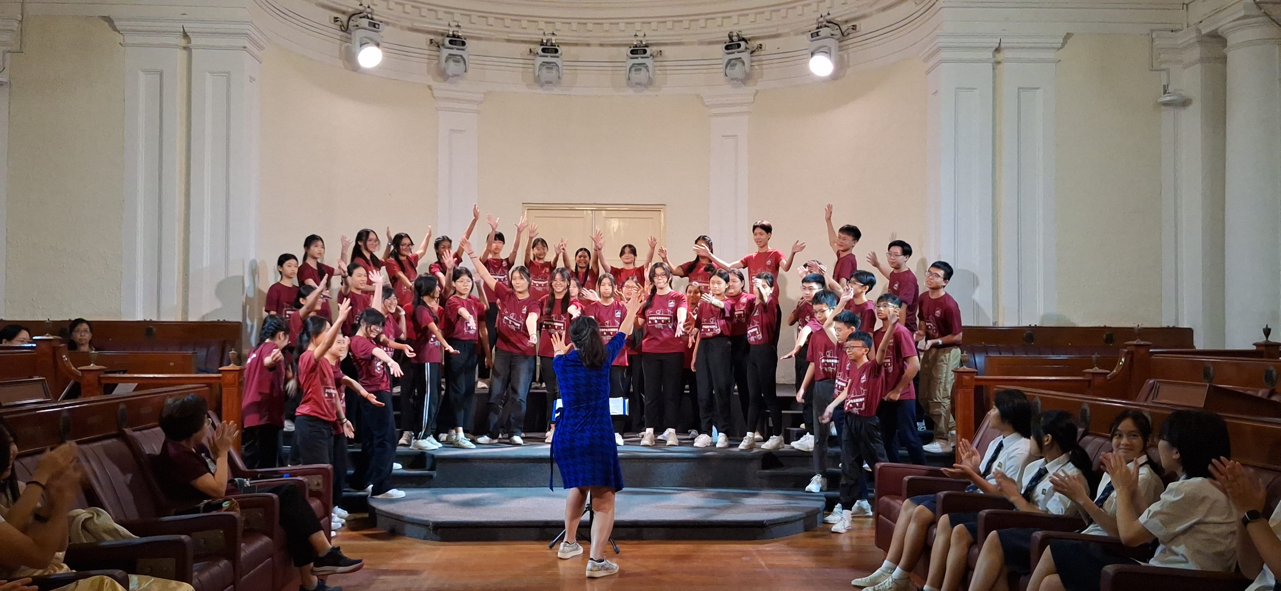 A conductor in blue leads a youth choir in red shirts performing on a stage in a grand hall.