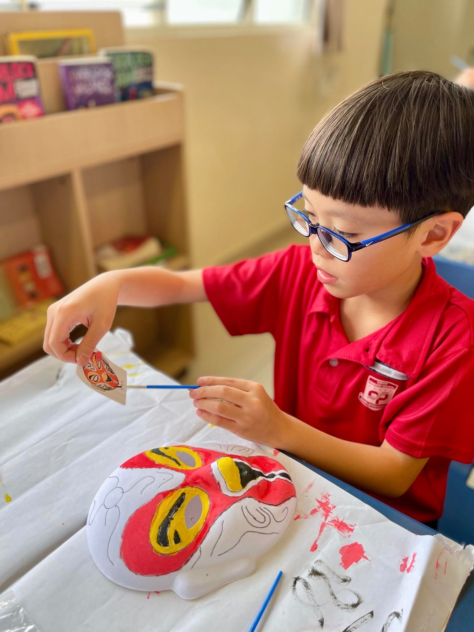 Young boy with blue glasses painting a colorful red and yellow traditional mask at a classroom table.