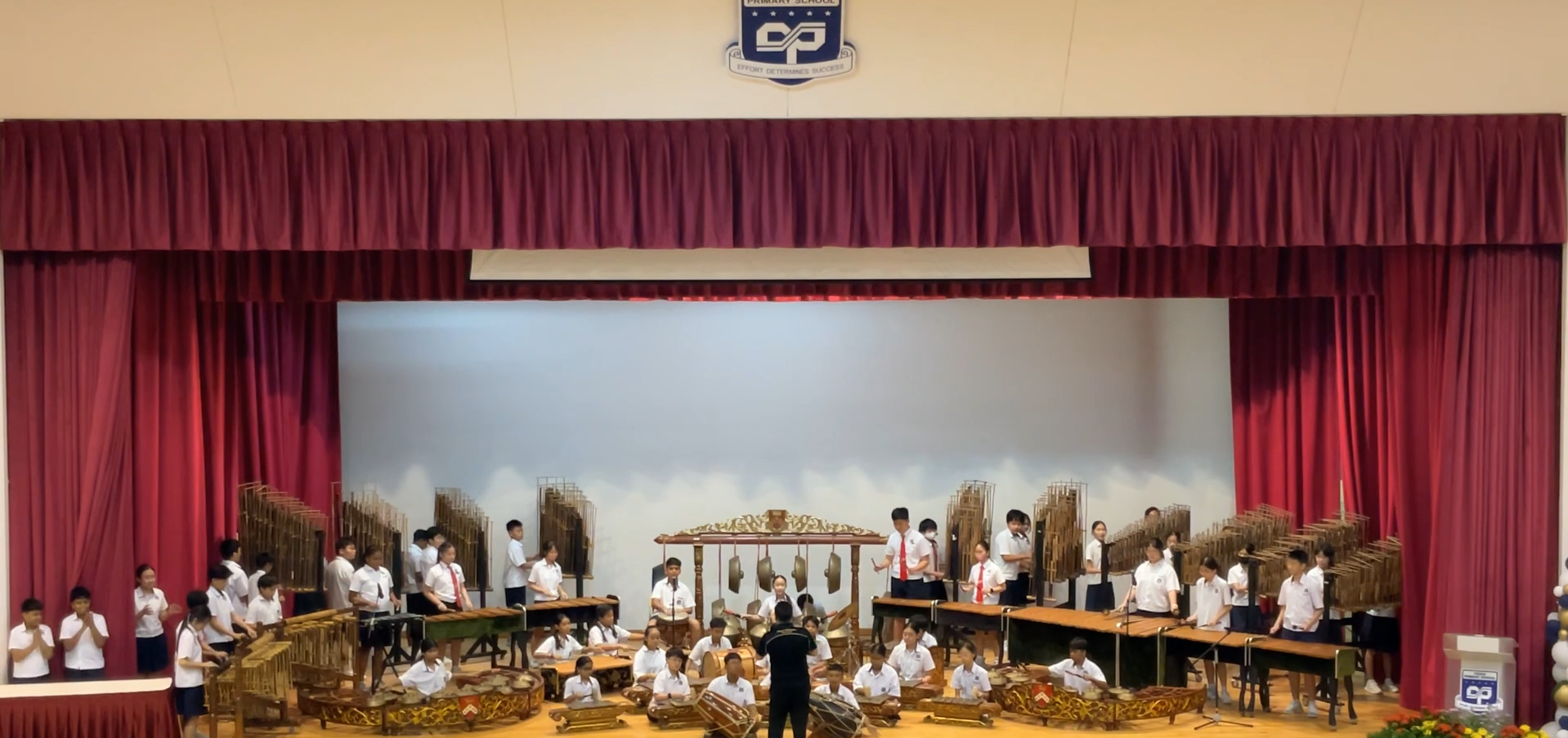Students in white uniforms perform a gamelan concert on a school stage with red curtains and a conductor.