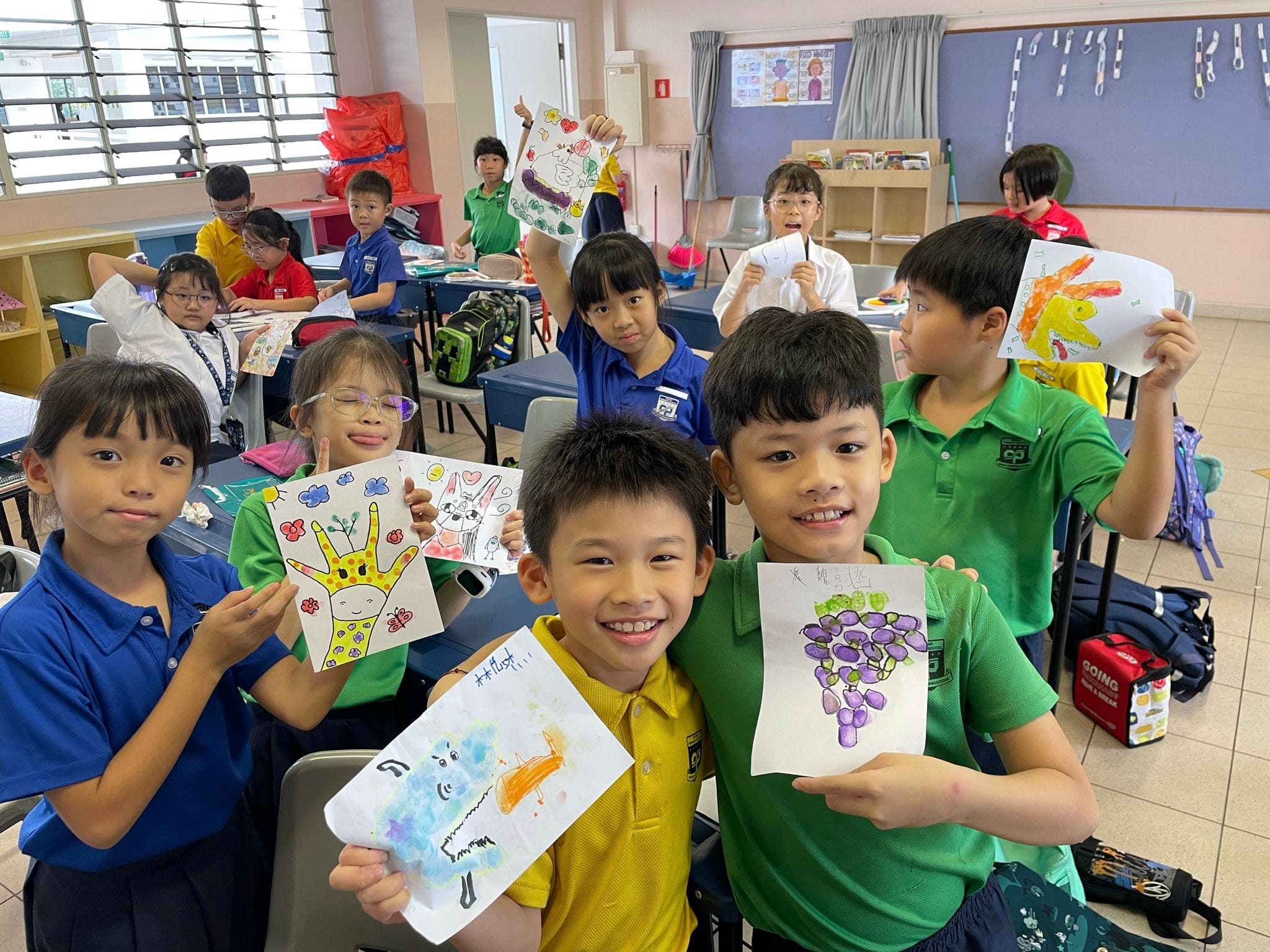 Young students in a classroom proudly hold up their colorful artwork including drawings of grapes, animals, and a hand.
