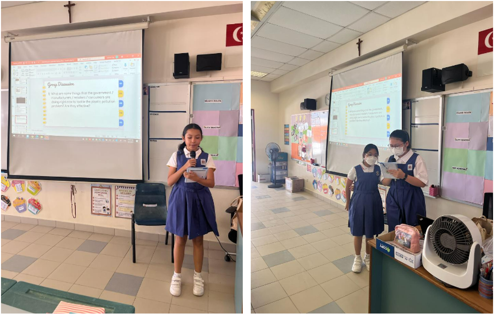 Two schoolgirls in navy uniforms present in a classroom with a projected slideshow on a screen behind them.
