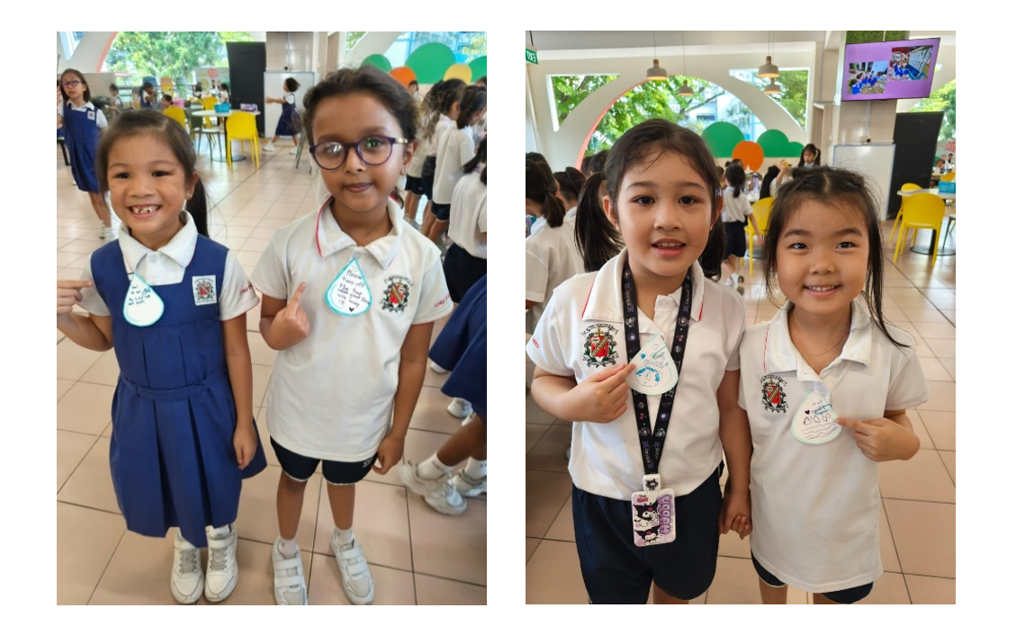 Four young schoolgirls in uniforms smiling and holding small decorated paper badges in a school cafeteria.