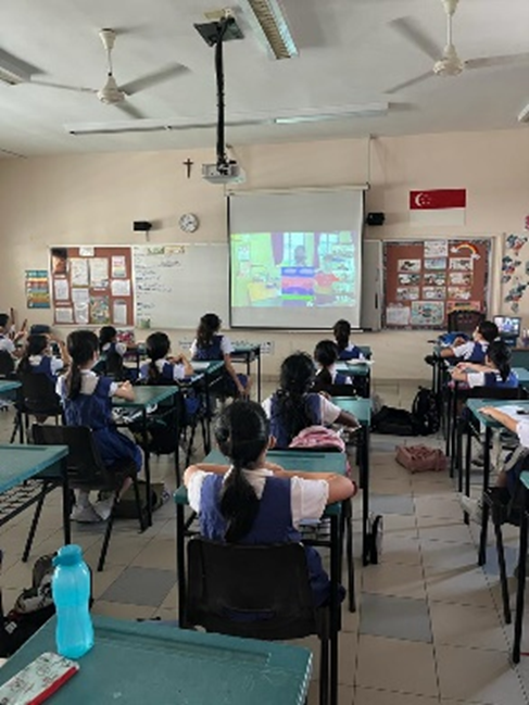 Students in school uniforms sit at desks watching a projected screen in a classroom with bulletin boards and ceiling fans.