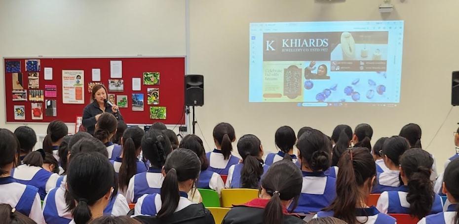 A woman speaks to a large group of students in blue vests, with a Khiards Jewellery website projected on screen.