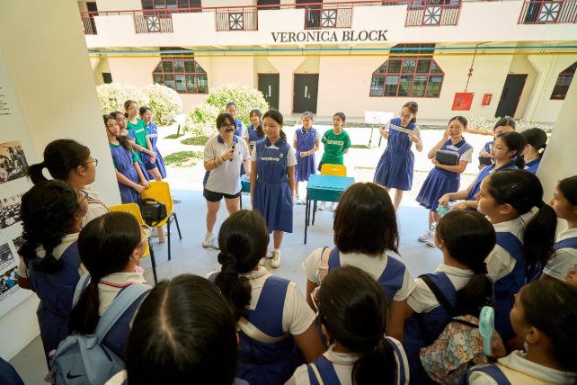 Students in blue uniforms gather in a circle outdoors near a building labeled Veronica Block.