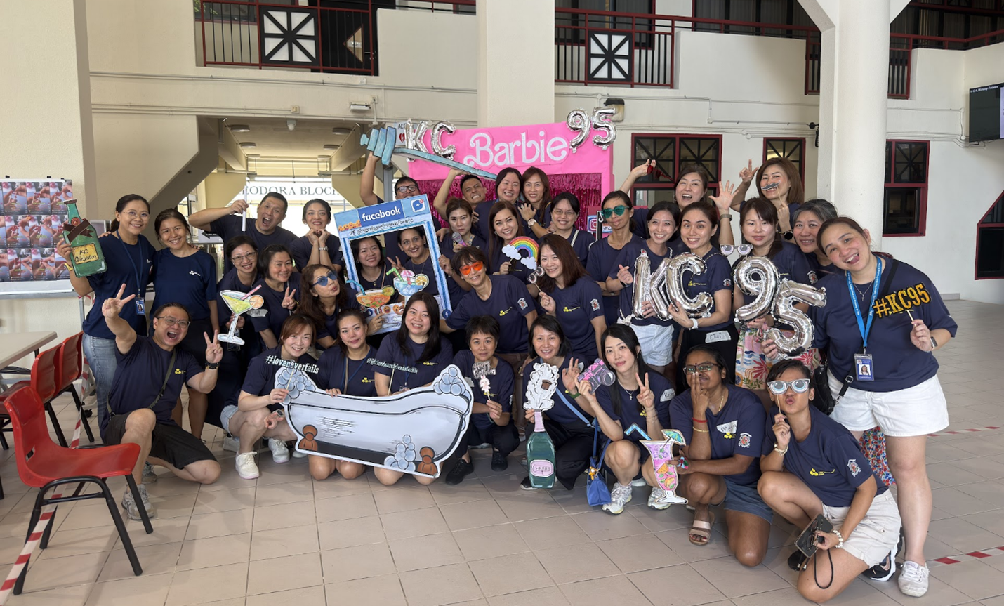 Large group in navy shirts posing with KC95 balloons, Barbie banner, and fun photo props indoors.